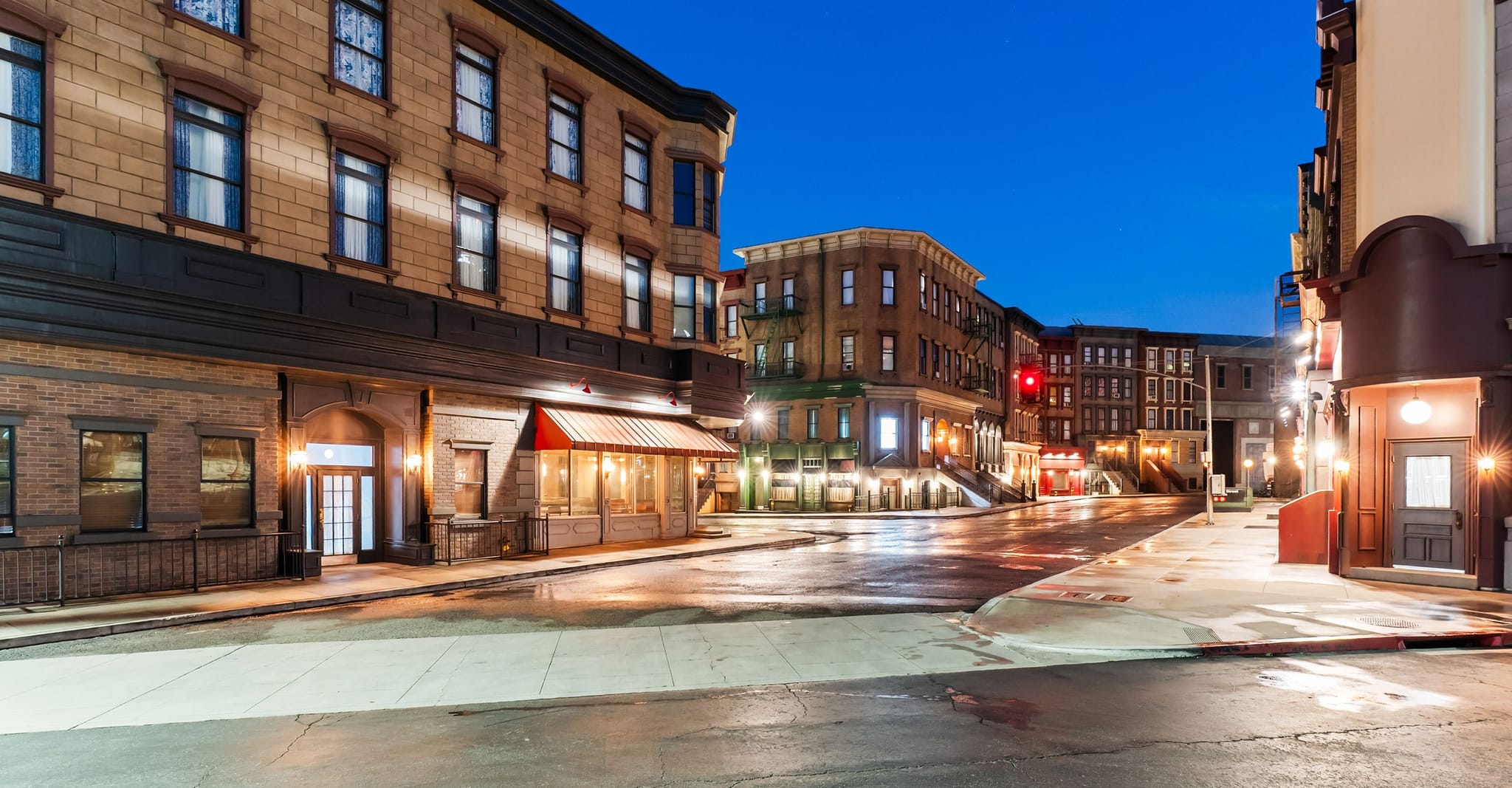 An empty, wet city street at dusk, reflecting lights from surrounding brick buildings.