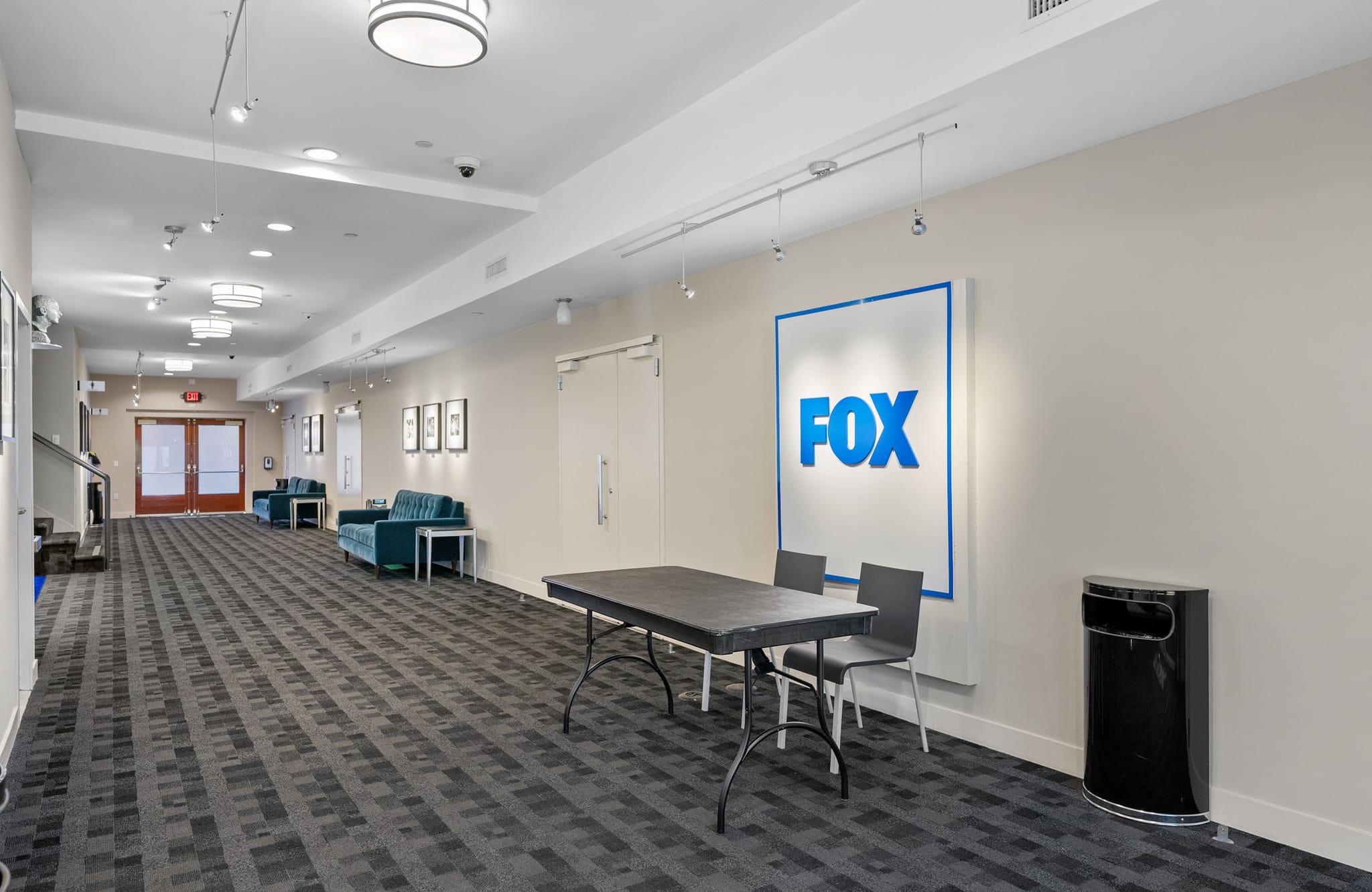 A long modern hallway with a large illuminated FOX logo sign, a black table with chairs, and teal couches.