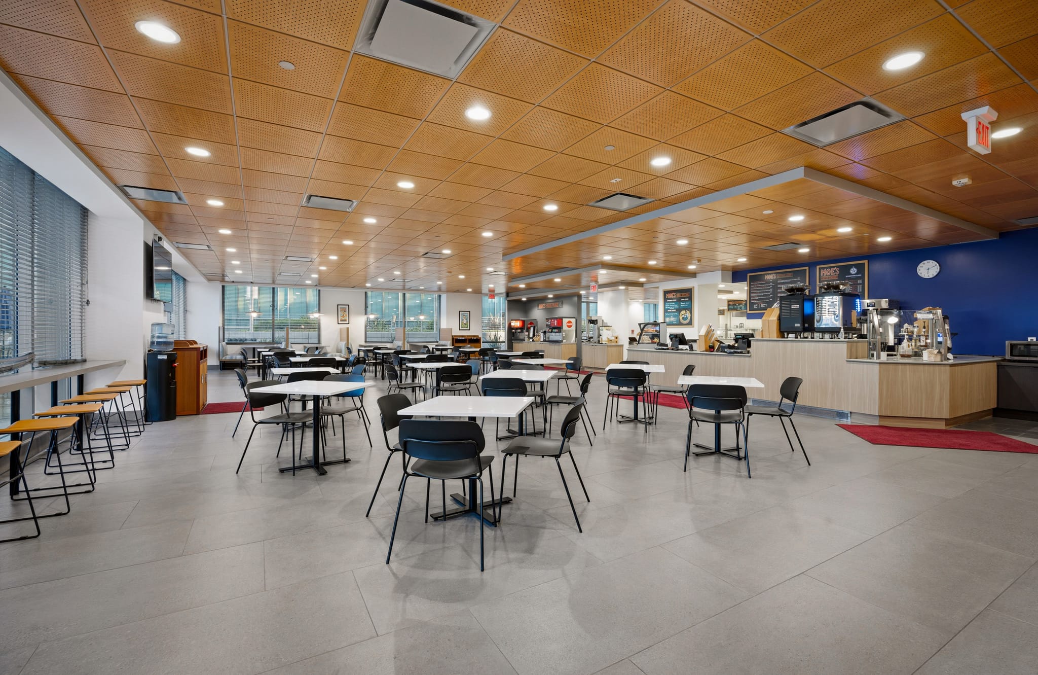 A modern, empty cafeteria with tables, chairs, windows, and a food service counter.