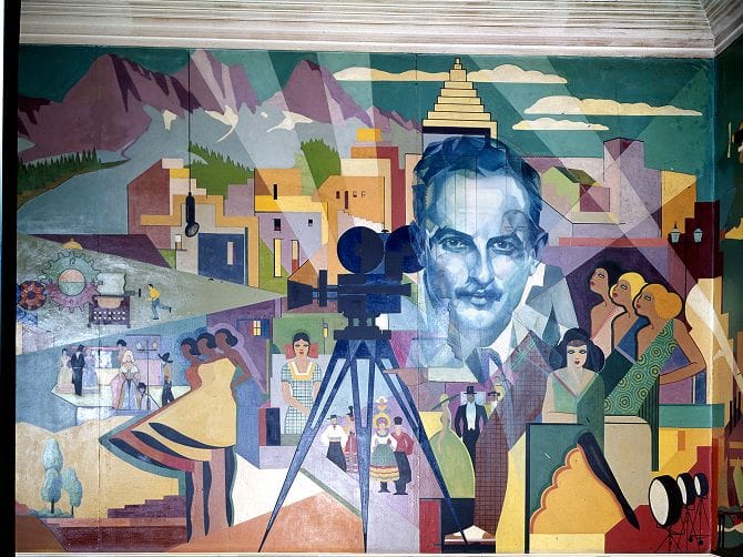 Vibrant Art Deco mural featuring a blue-toned portrait of a man, a film camera, and diverse scenes of people against a stylized city and mountains.