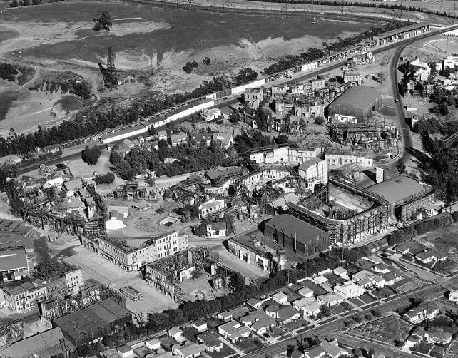 Aerial black and white view of a large film studio complex with multiple sound stages, extensive backlot sets, and construction, bordered by residential homes and hills.