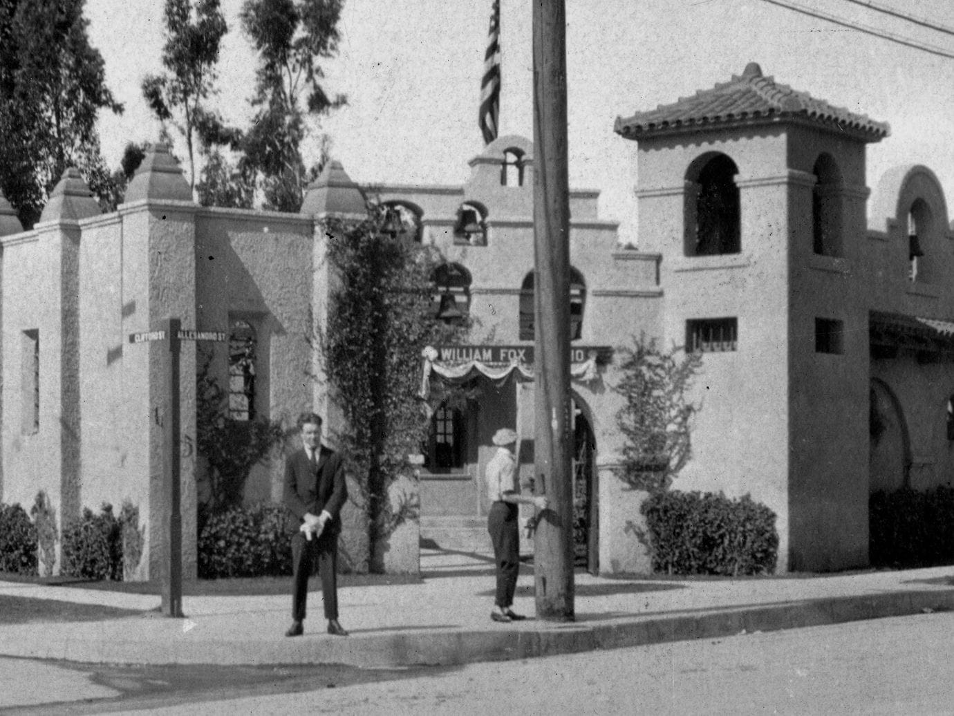 Black and white photo of the Spanish Revival style William Fox Studio building with a tower and American flag, and two men on the sidewalk.