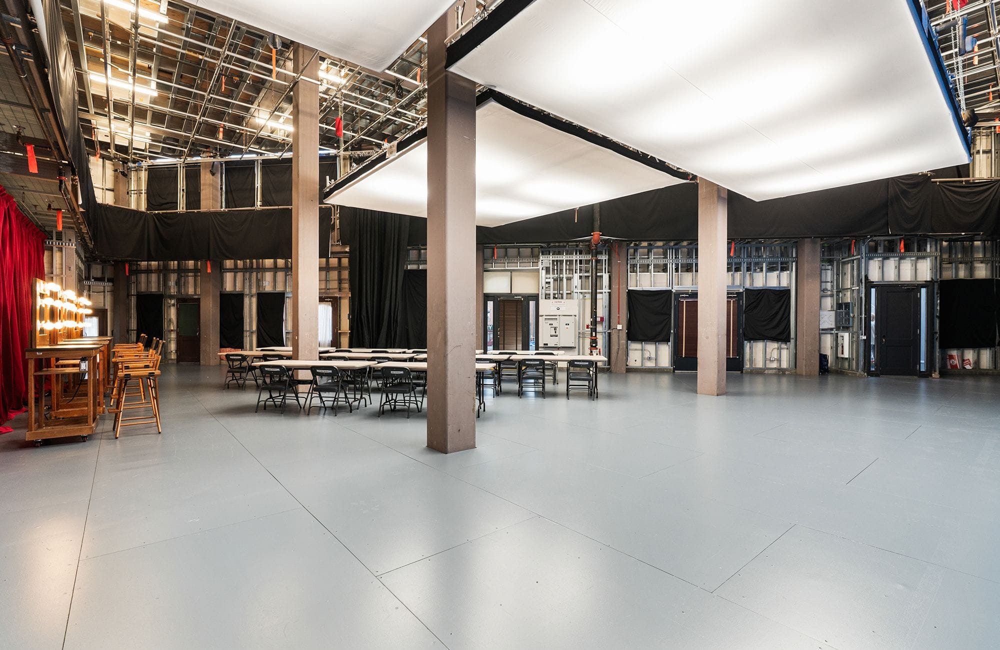 A spacious studio with an exposed ceiling, brown pillars, tables and chairs, and a lighted makeup vanity.