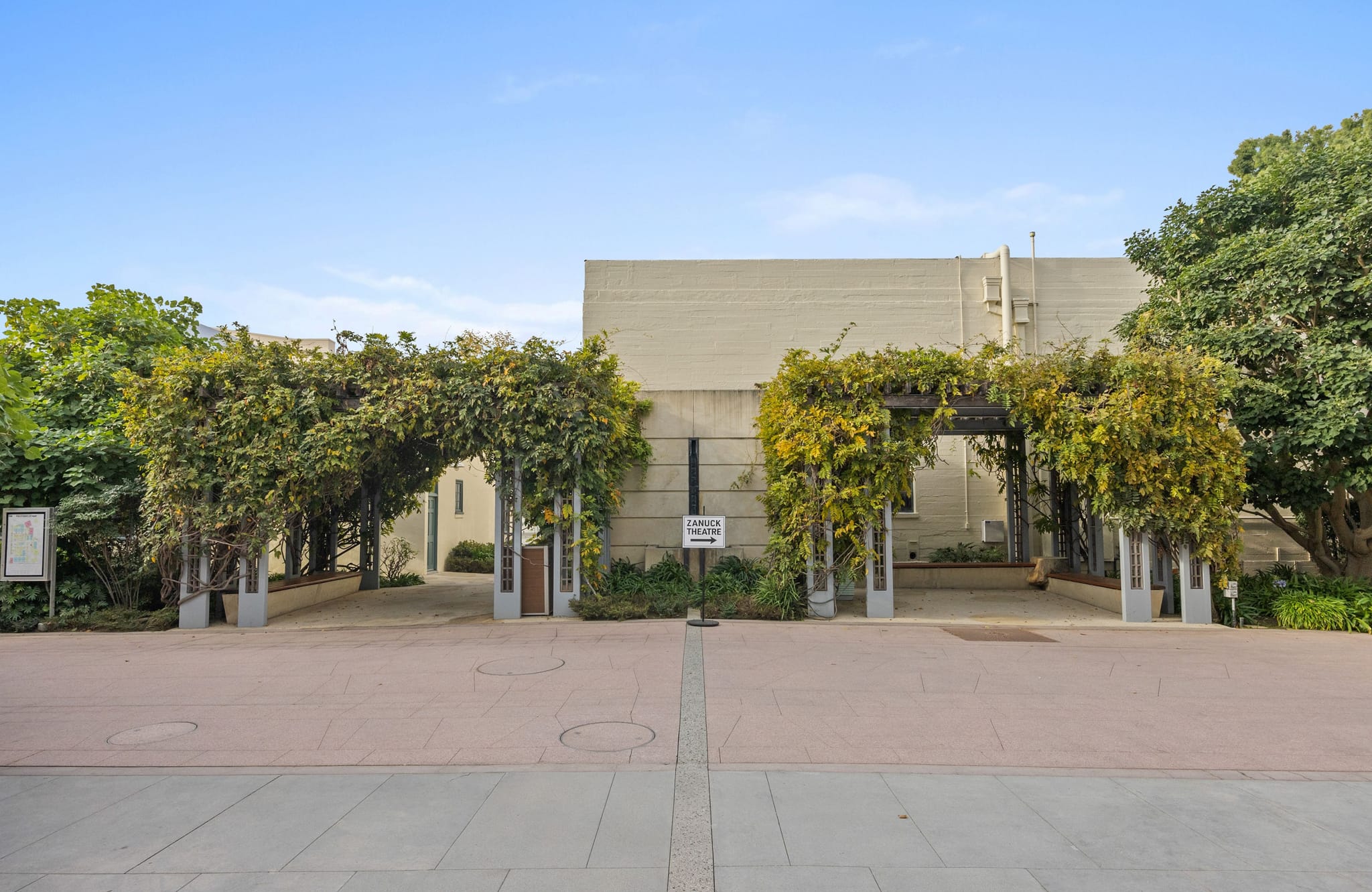 A paved path leads to a building entrance through a lush, vine-covered arbor, with a sign for Theatre Guild on the right.