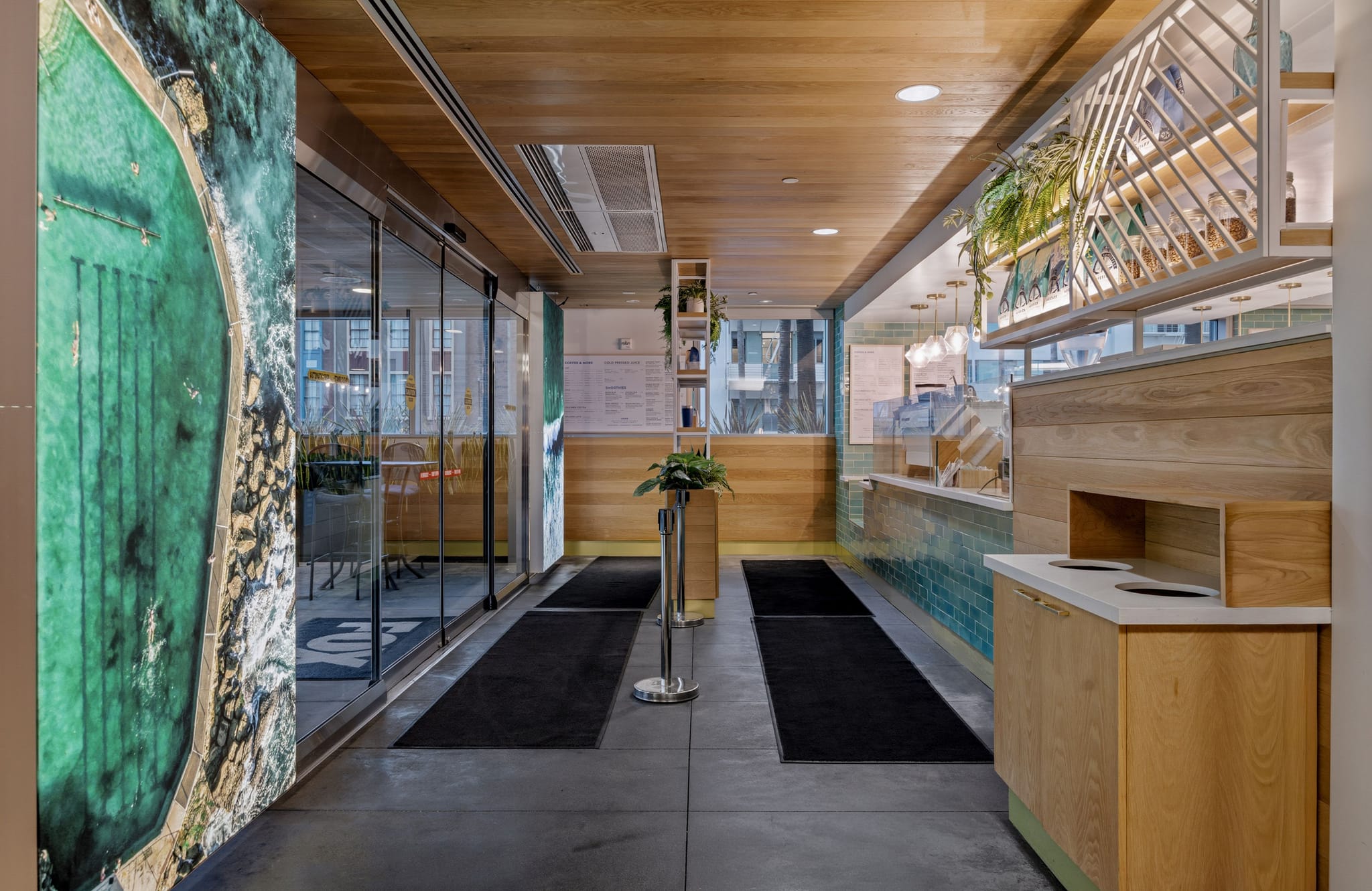 A modern cafe or restaurant entryway with a large mural of a sea pool, glass doors, and a service counter with wooden and tiled accents.