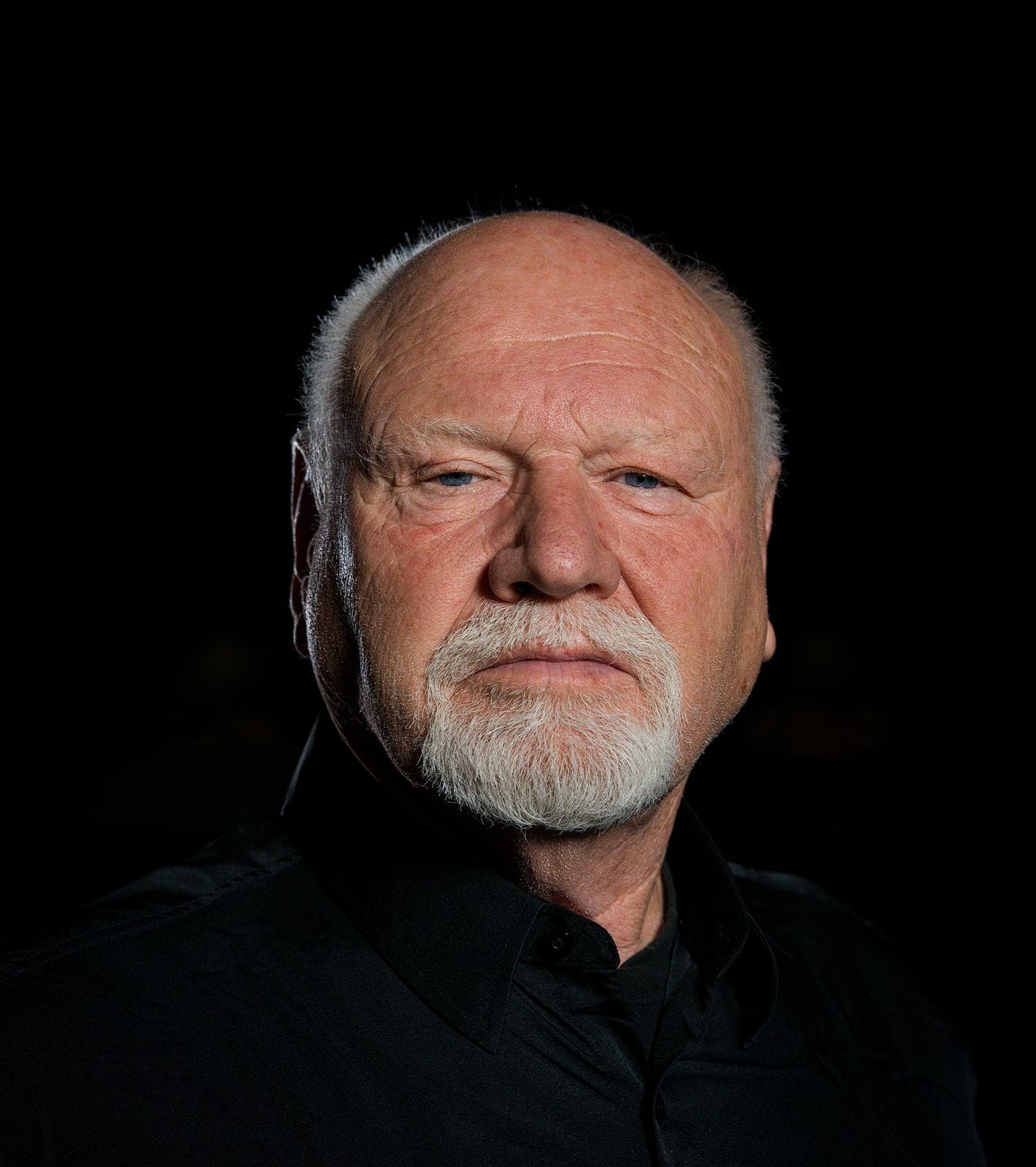 Headshot of a bald man with a white beard and a serious expression, wearing a black shirt.