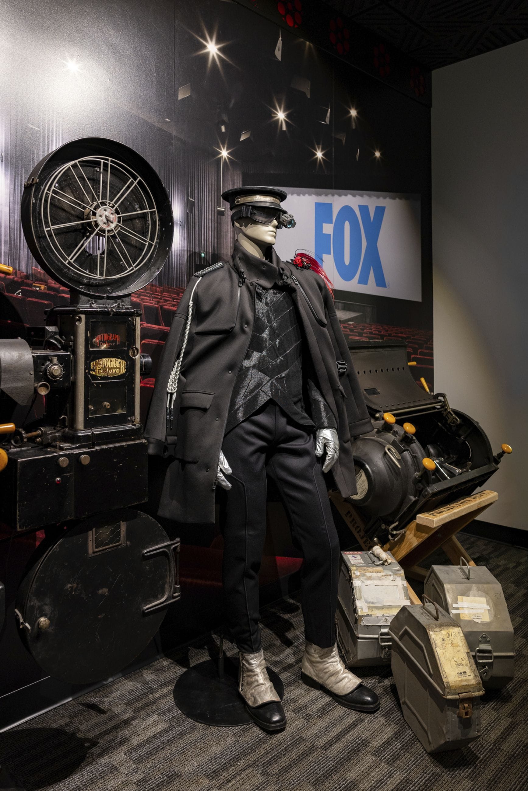 A mannequin in a dark uniform with goggles and silver accents stands between vintage film projectors, with a movie theater backdrop showing "FOX".