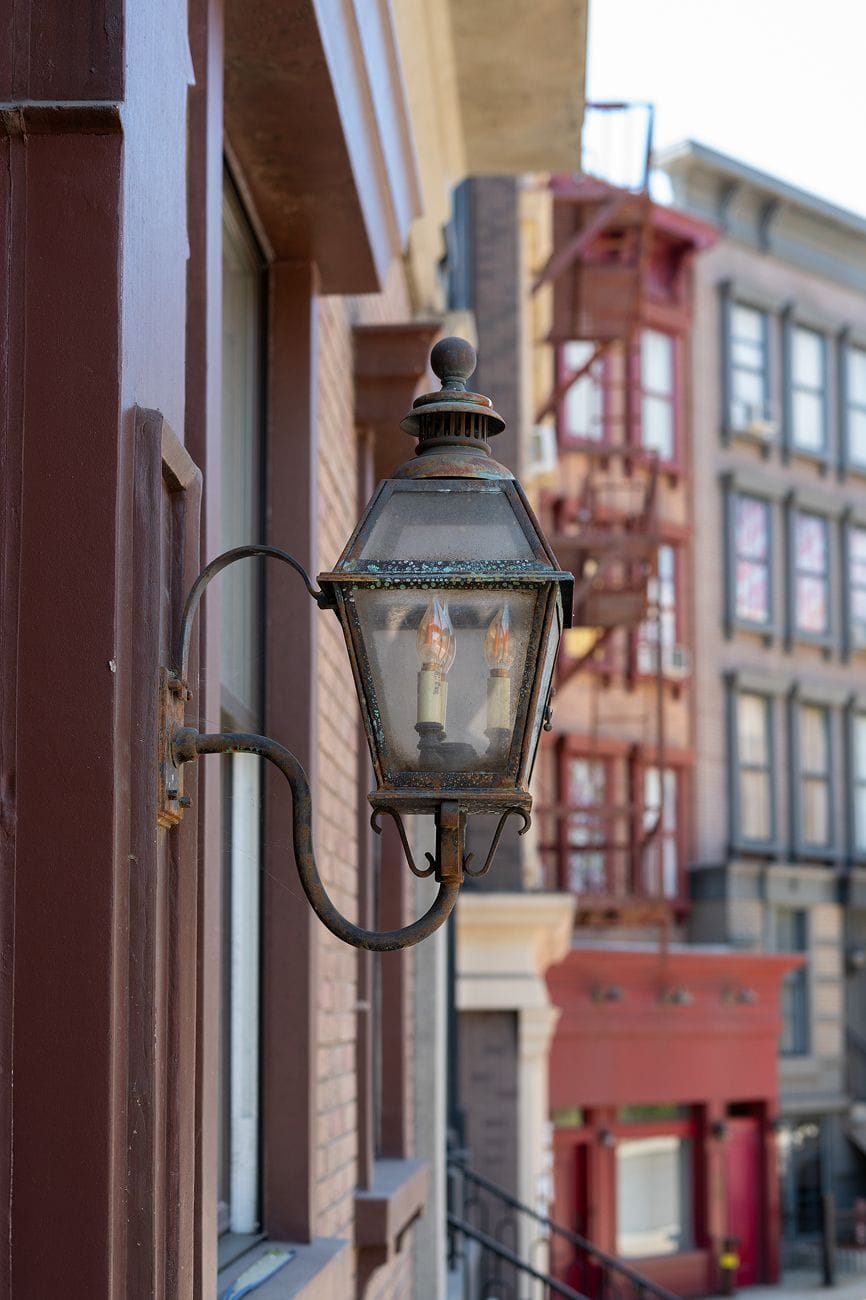 A rusty old-fashioned lantern with two flame-shaped bulbs on a brown building, with other buildings blurred in the background.