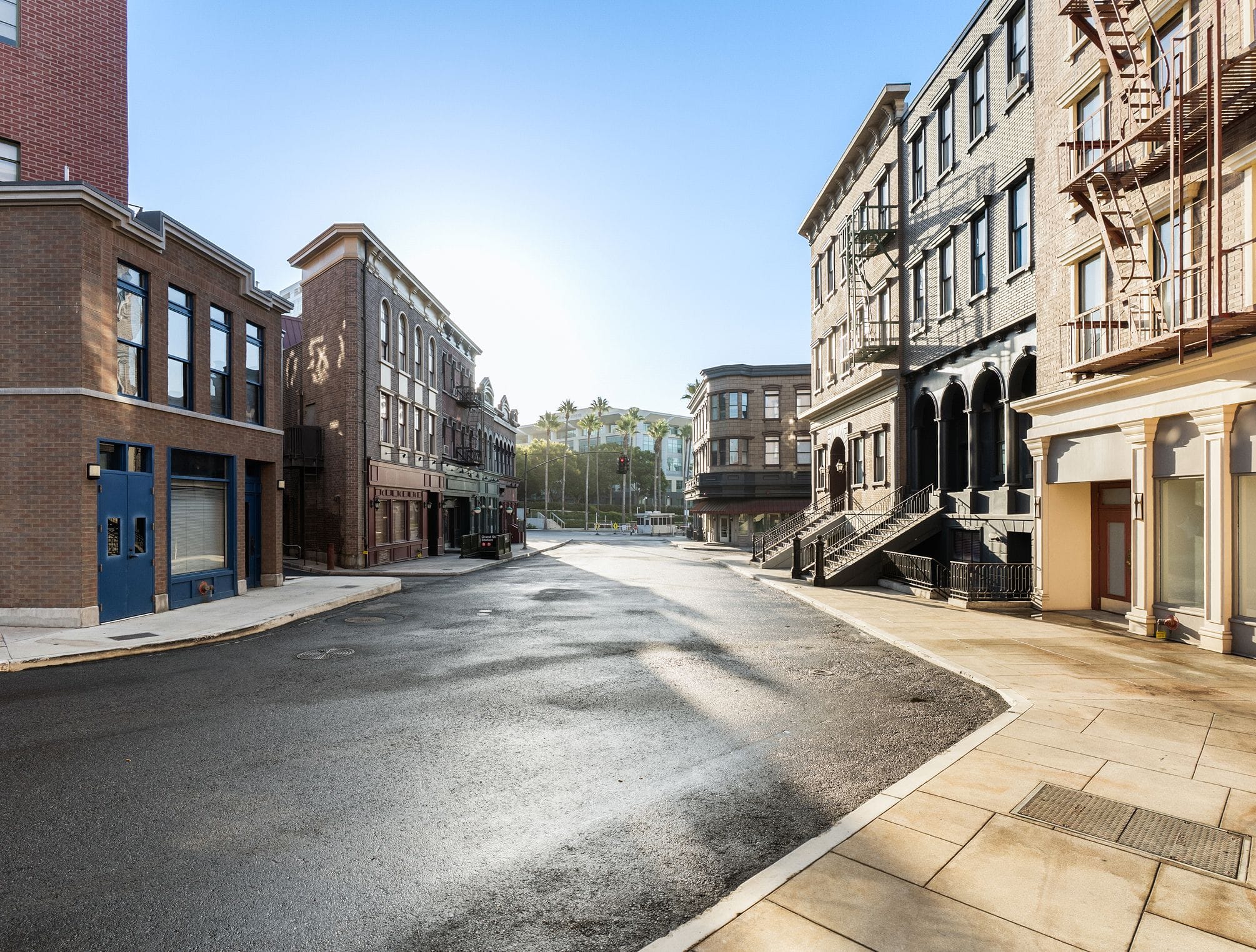Empty street lined with multi-story brick buildings with fire escapes under a clear, sunny sky.
