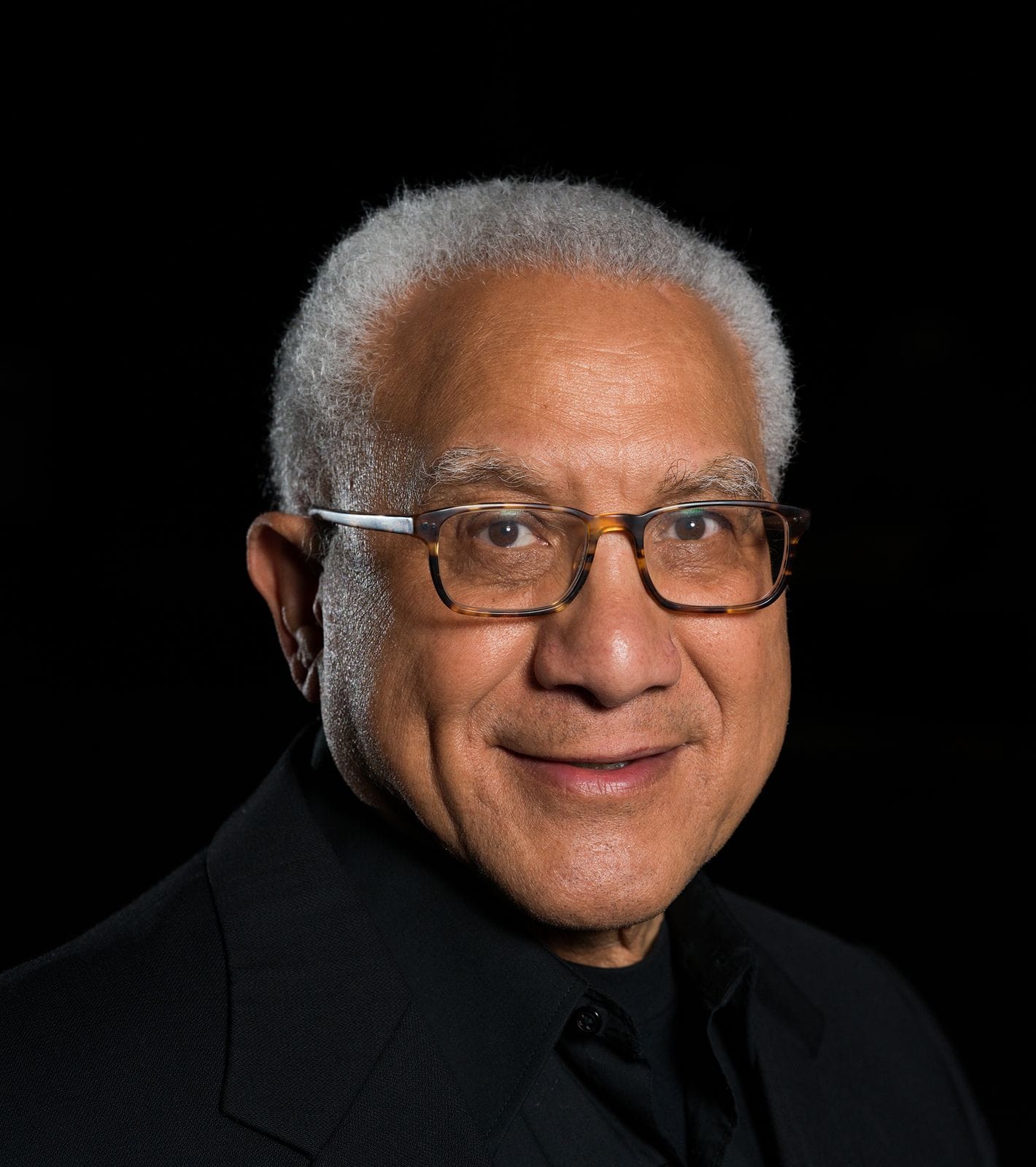 Smiling older man with gray hair and glasses against a black background.