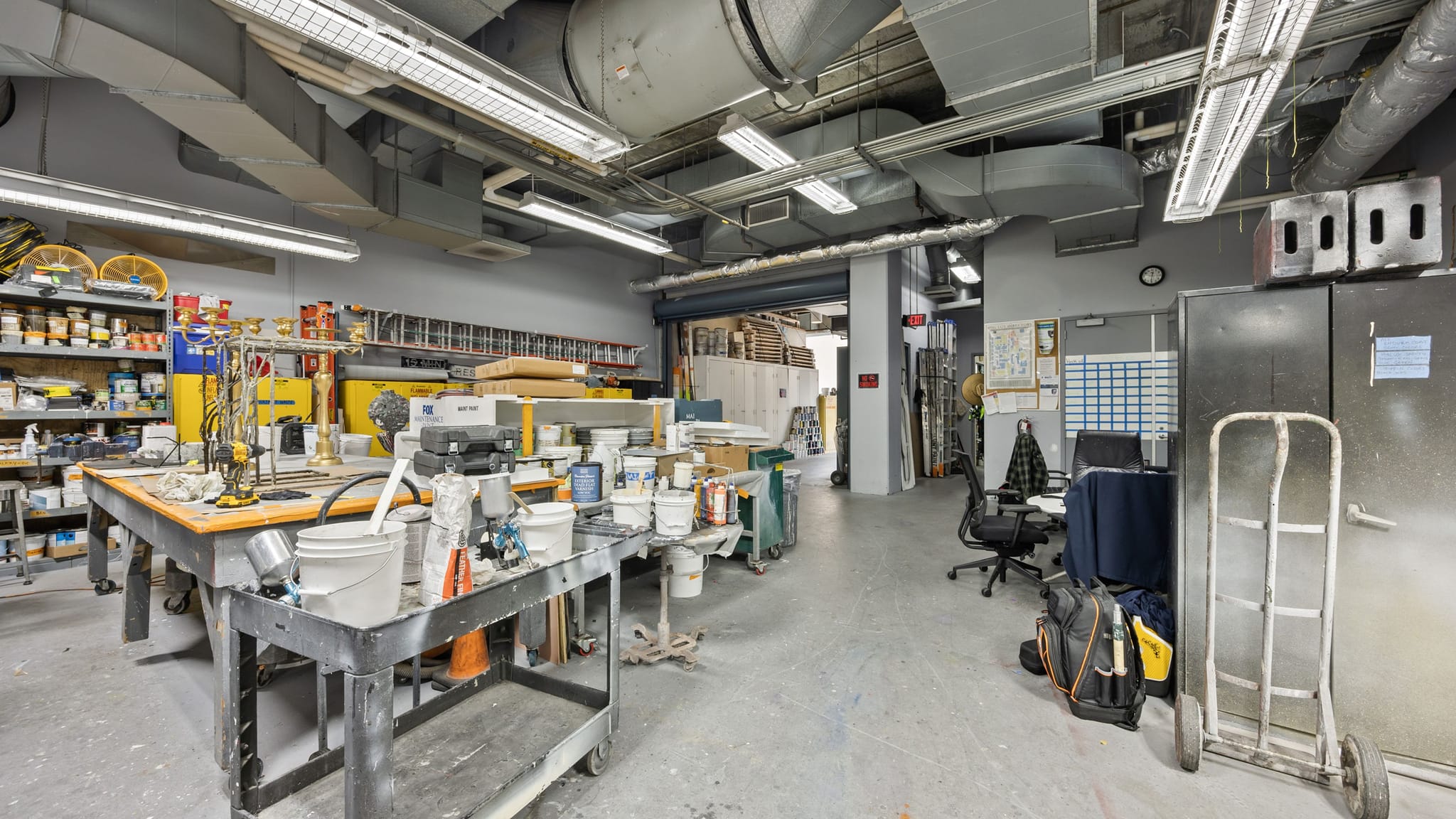 Industrial workshop with exposed ceiling ducts, workbenches, paint supplies, and an office area.