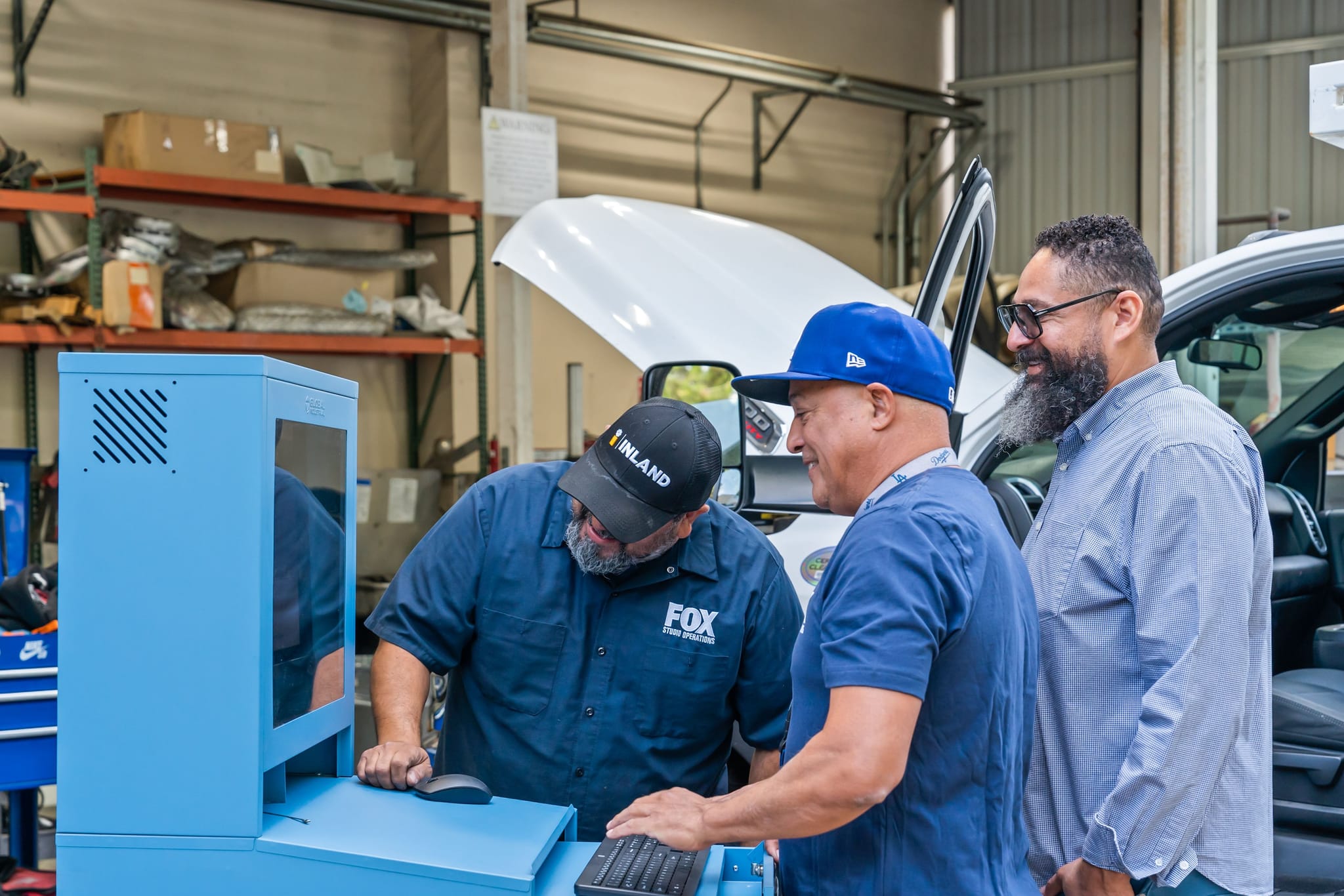 Three smiling men work on a blue diagnostic machine in a garage with a white truck.