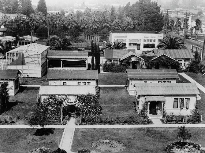 Elevated black and white view of bungalows, green spaces, and a rose-covered arch, with a "MOTOR SUPPLY" building behind.