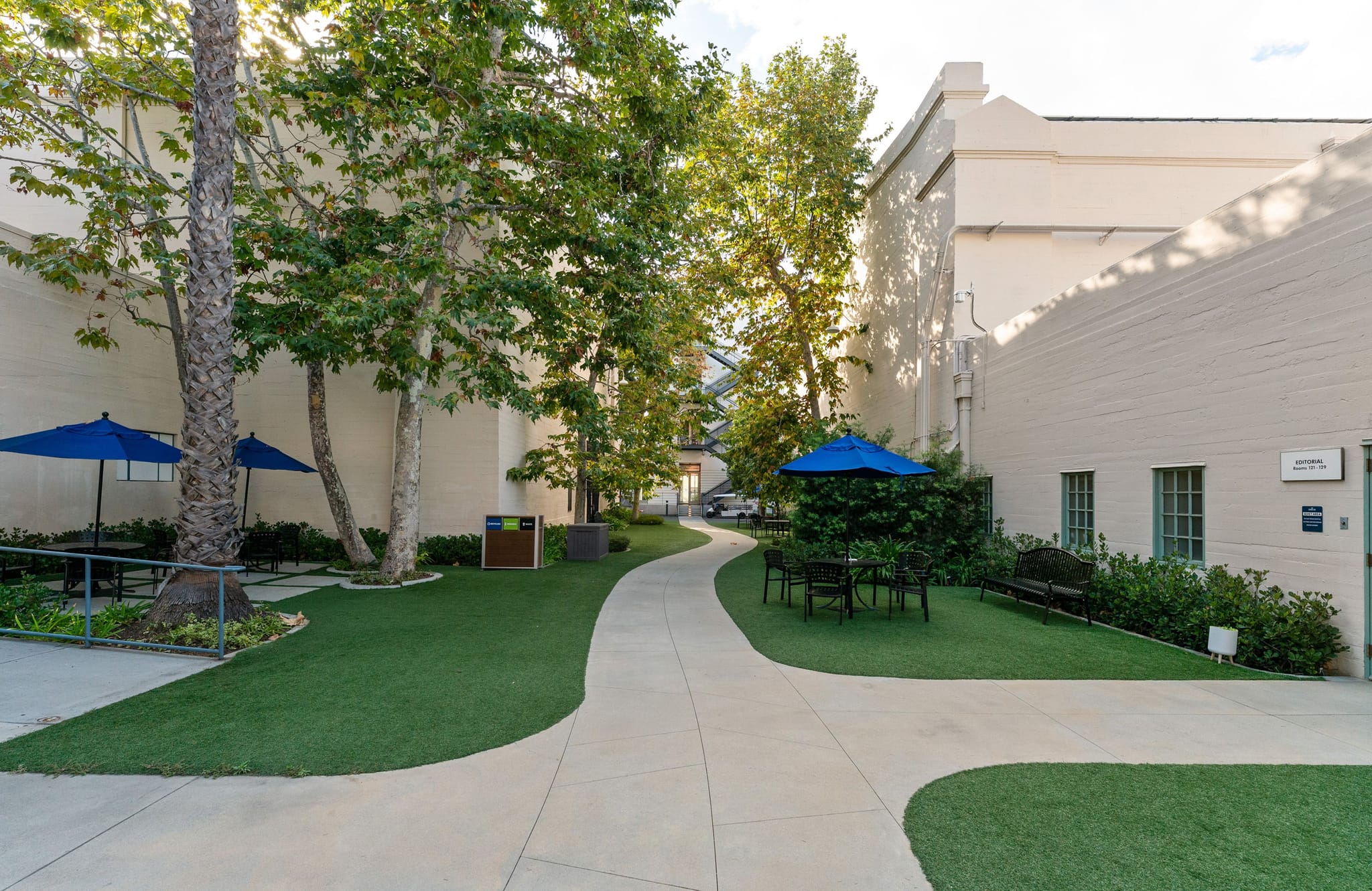 A green courtyard with a winding path, trees, outdoor seating with blue umbrellas, and light-colored buildings.