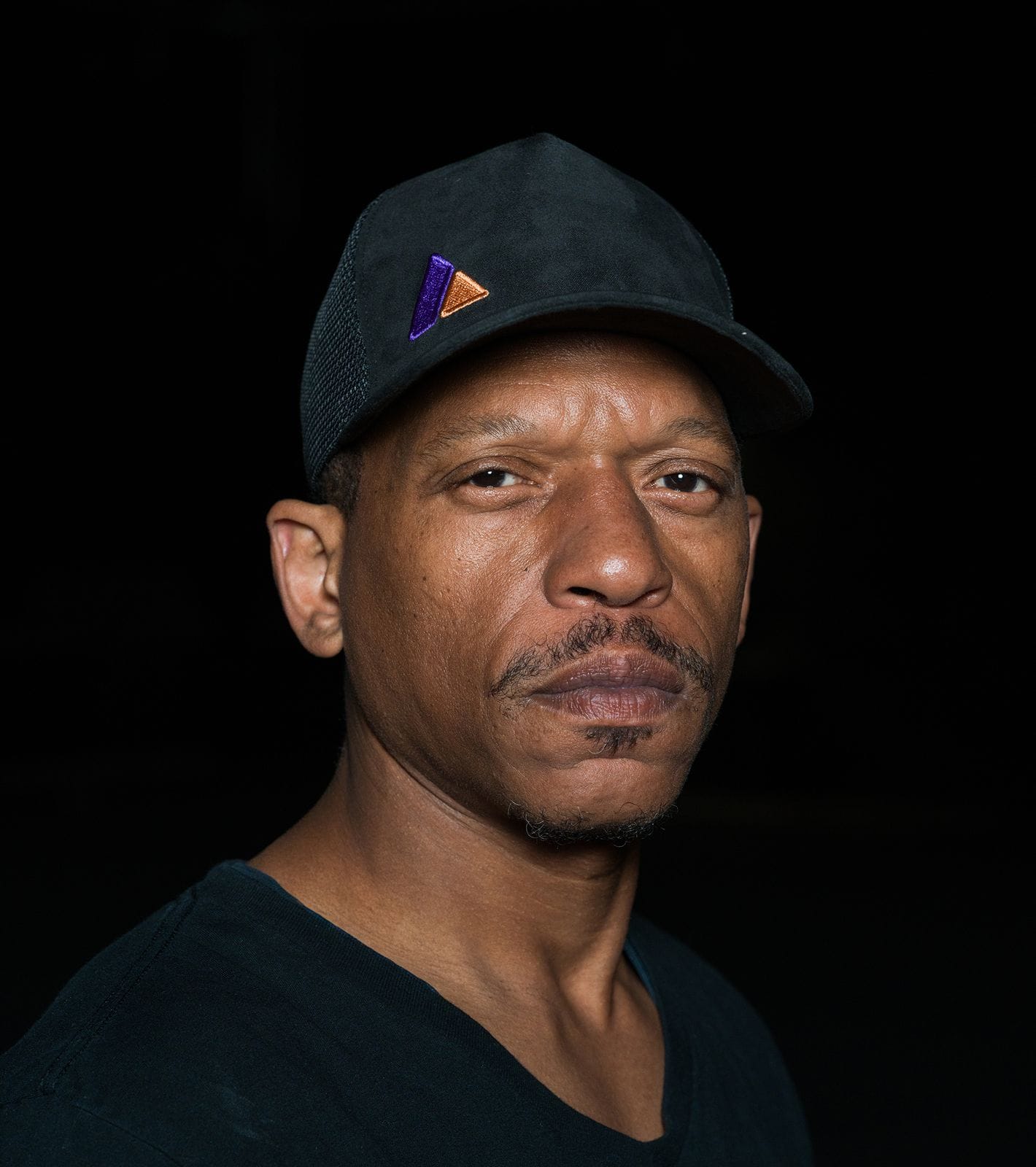 A man with a mustache and soul patch wears a black cap with a purple and orange logo and a black shirt, looking directly forward against a dark background.
