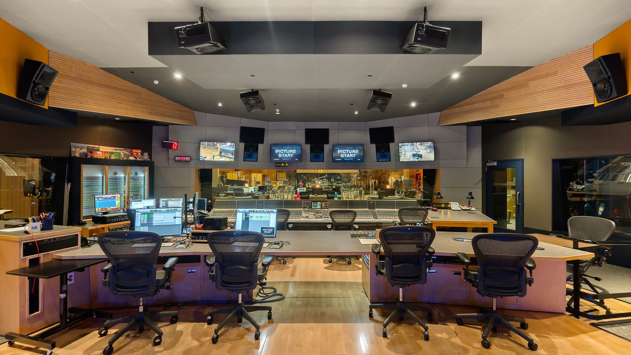 A modern audio/video production control room with multiple workstations, a large mixing console, and monitors showing 'PICTURE START'.