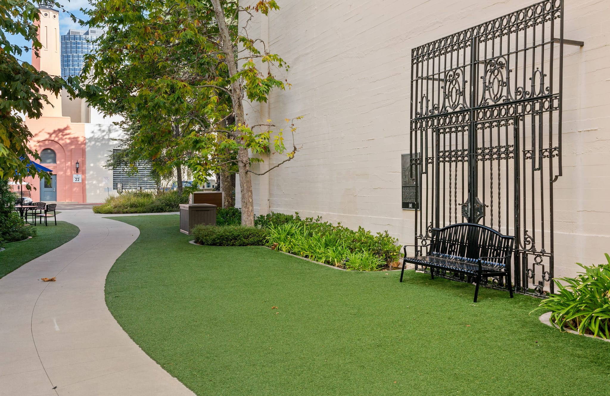 A curved pathway through a green urban courtyard with a large decorative black gate and bench against a wall on the right.