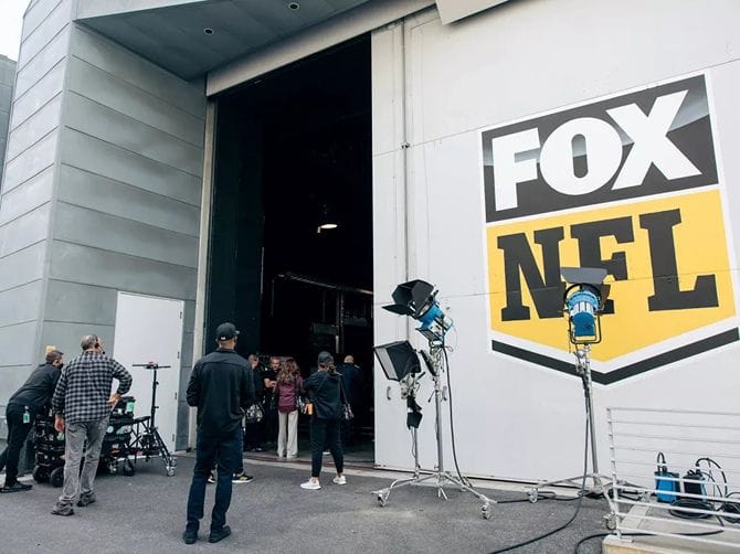 A production crew and lighting equipment are set up outside a building with a large FOX NFL logo.
