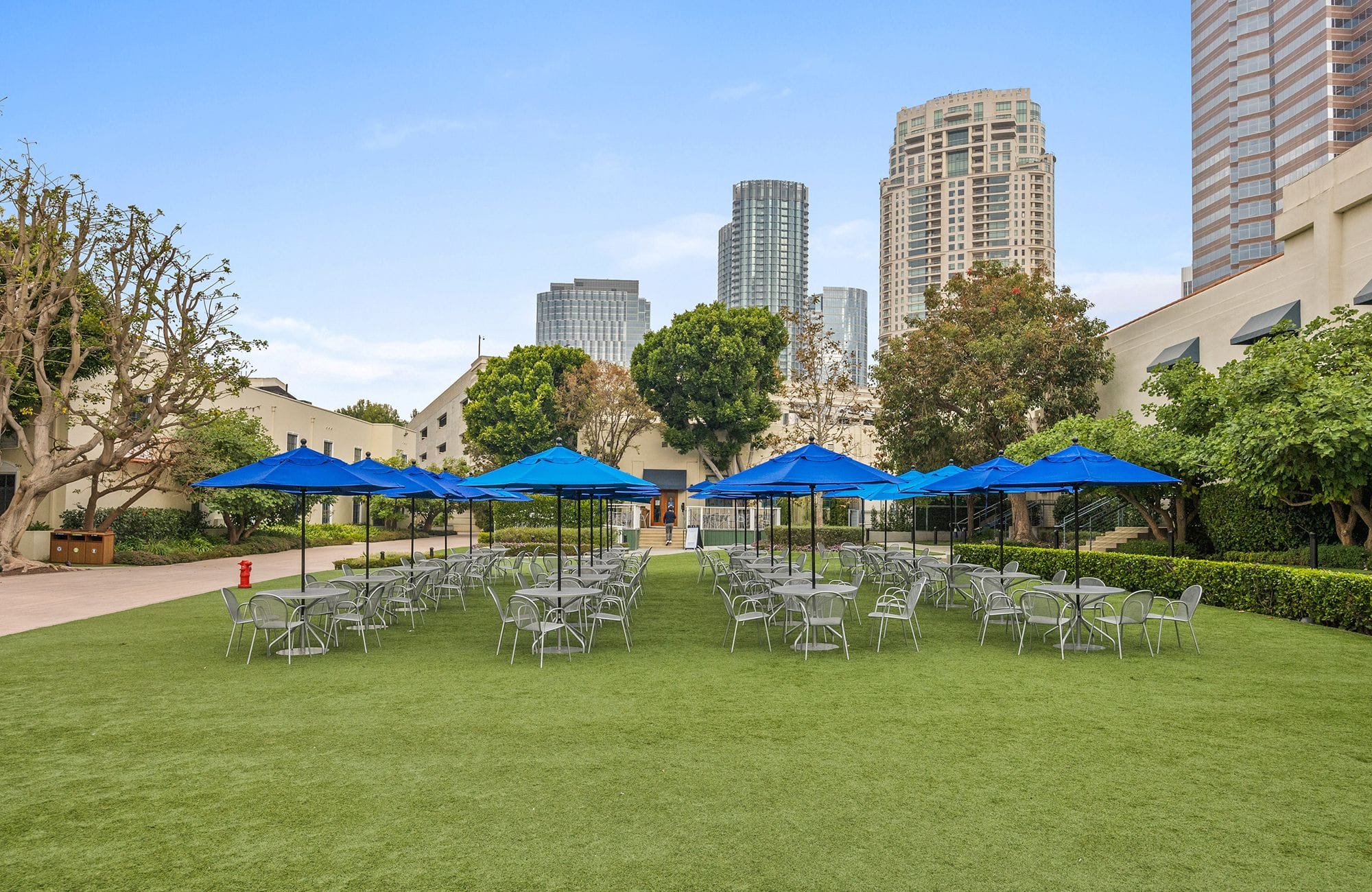 Outdoor seating on a green lawn with blue umbrellas, surrounded by buildings and skyscrapers.