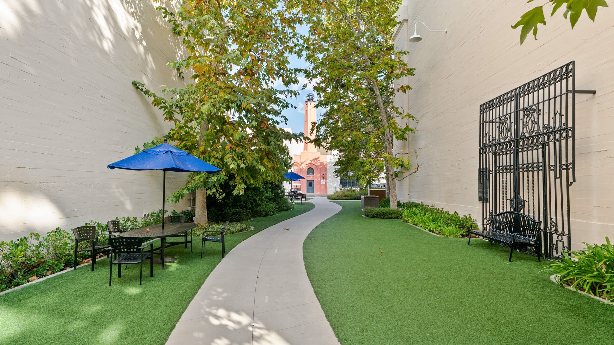 An outdoor area with a winding path through green turf, flanked by walls and trees, featuring seating, a decorative gate, and a distant brick building.