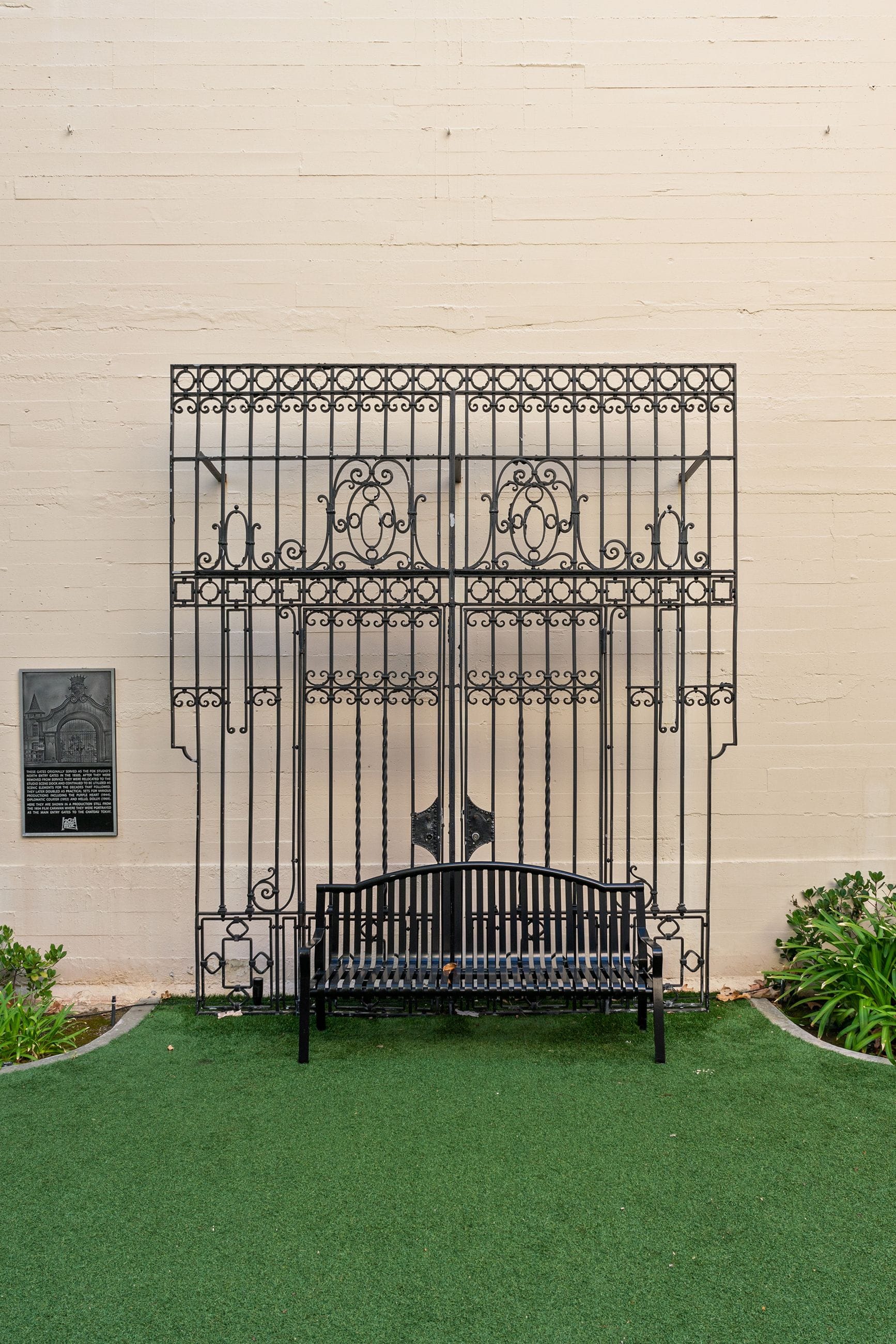 A black ornate wrought iron structure and matching bench sit on green turf against a cream wall.