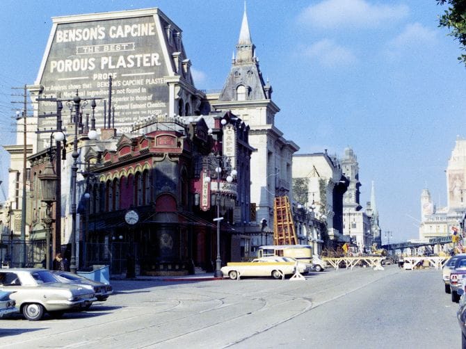 A vintage street view featuring ornate buildings, including one with a large "Benson's Capcine Porous Plaster" billboard, classic cars, and trolley tracks under a blue sky.