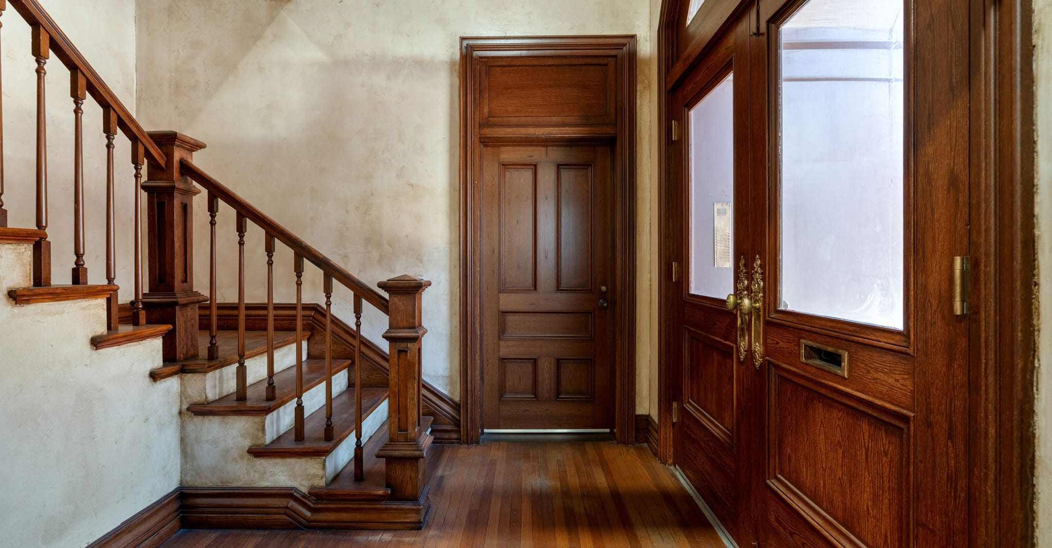 Interior hallway with a dark wood staircase, hardwood floor, and two wooden doors.