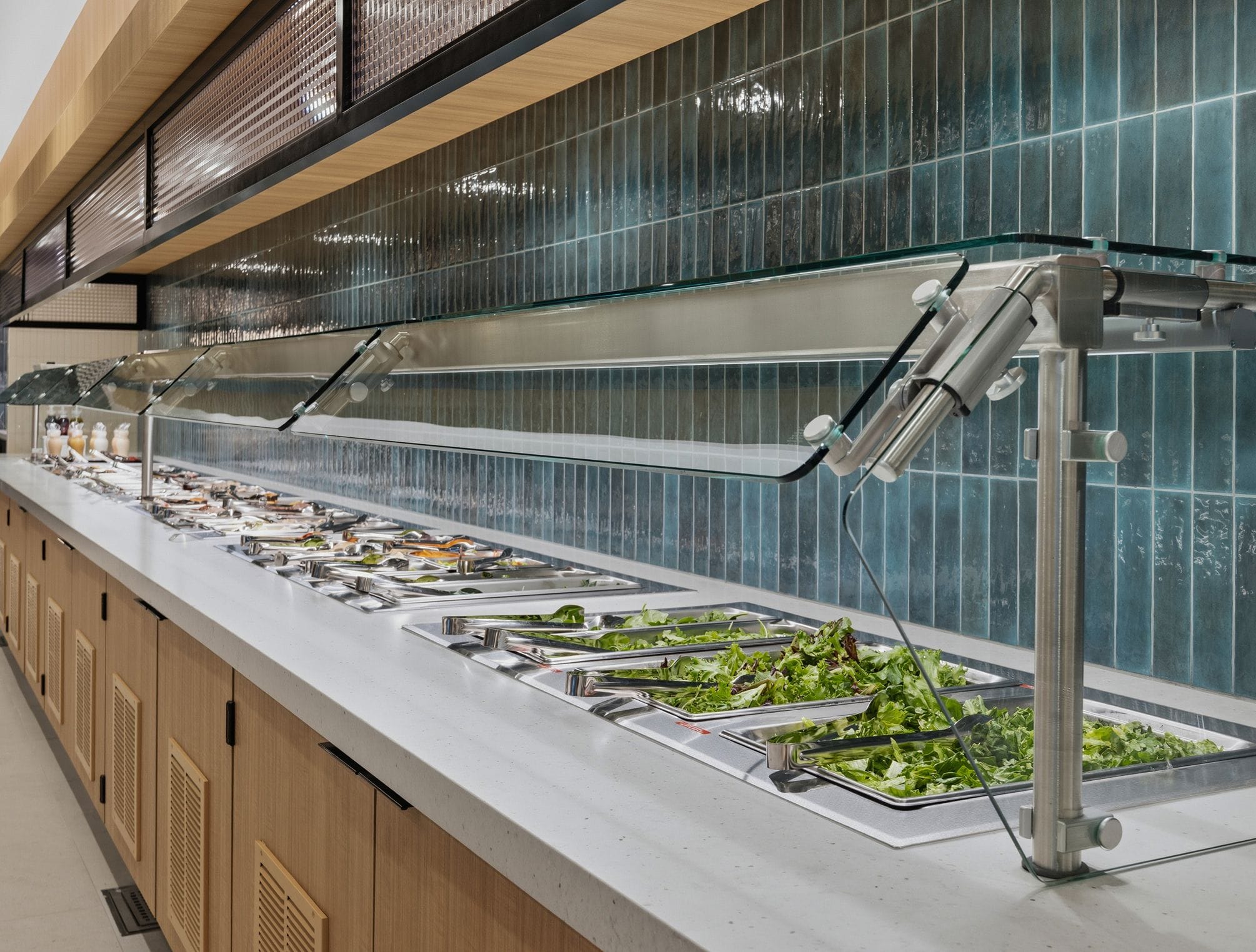 A modern self-service salad bar with fresh greens and other dishes under a glass sneeze guard, against a teal tiled wall.