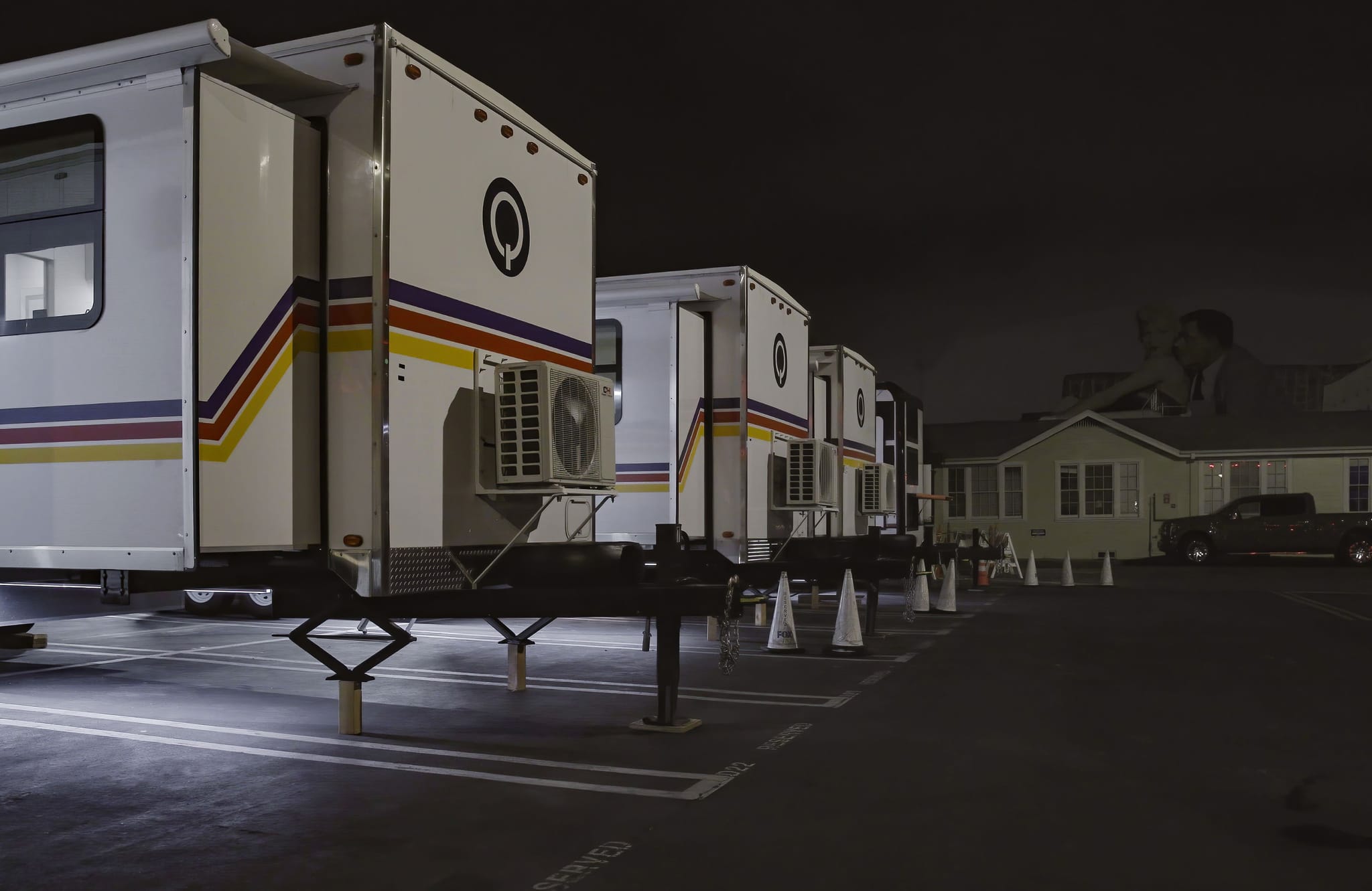 White mobile trailers with colorful stripes and AC units are parked at night, with buildings and a large, faint image of two men in the background.