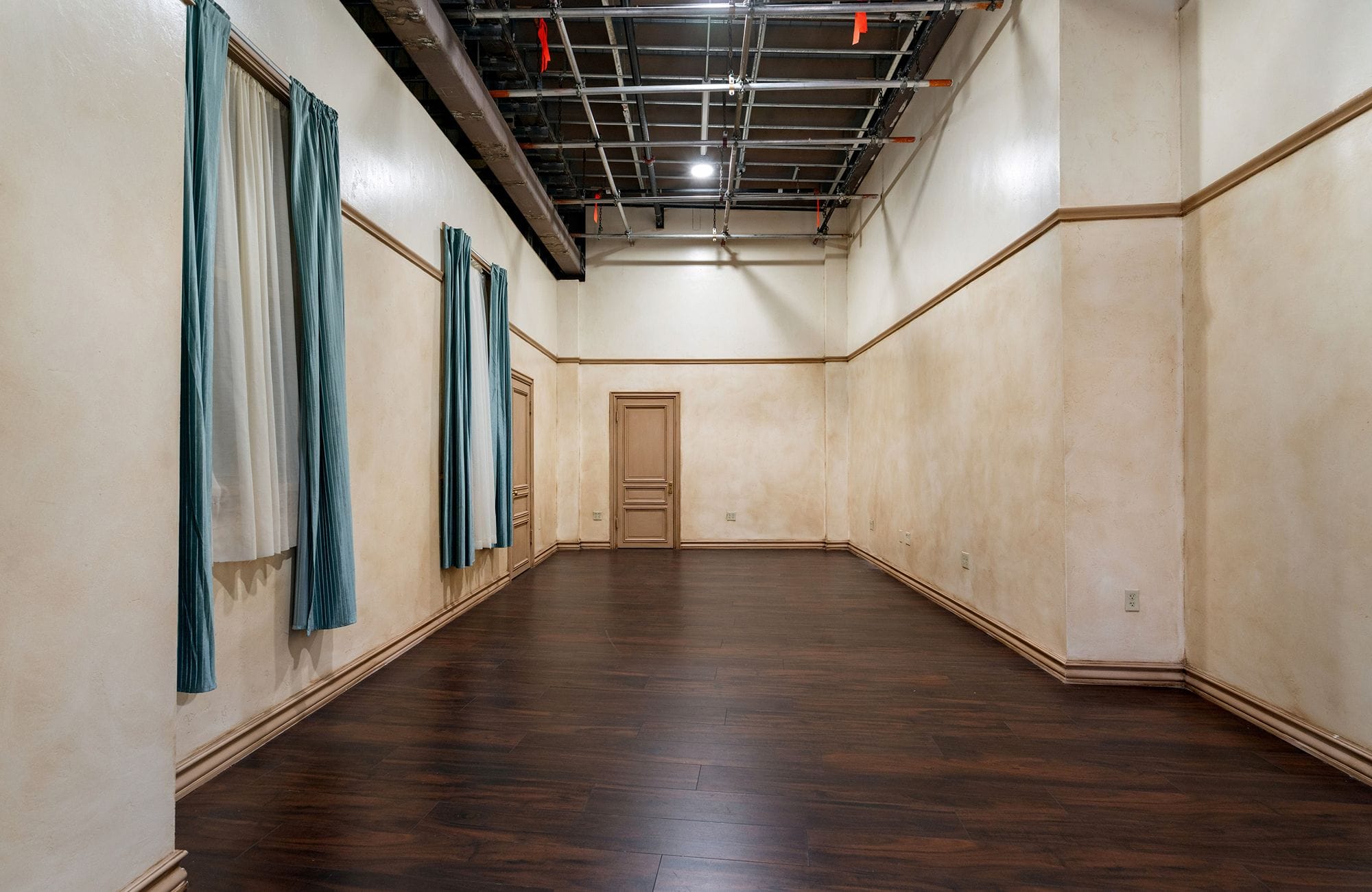 Empty room with dark wood floor, two-tone walls, blue curtains, two wooden doors, and an exposed ceiling grid.
