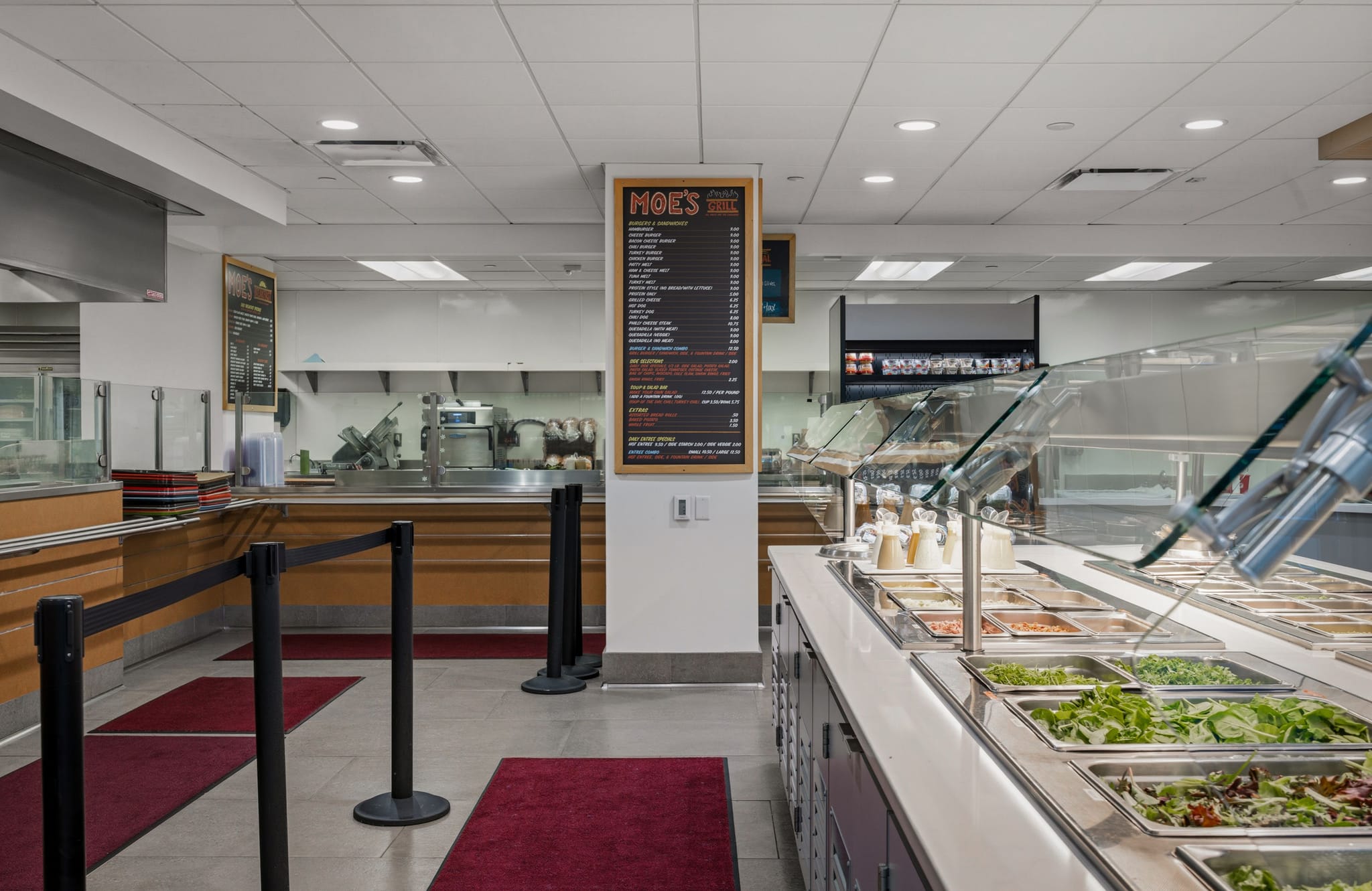 Interior of a cafeteria with a salad bar, a "Moe's" menu board, and a food service counter in the background.