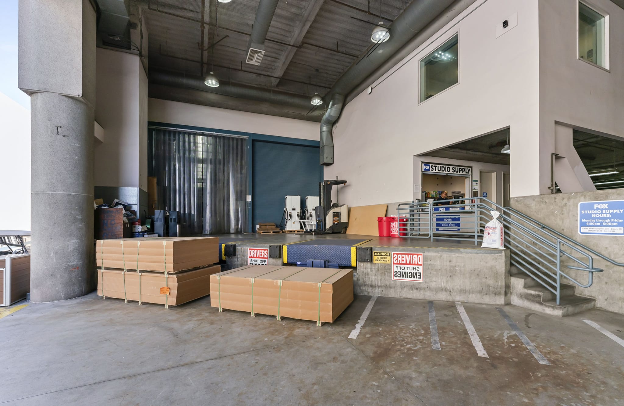 An industrial loading dock area with stacks of packages on the concrete floor, a raised platform leading to "Studio Supply," and a staircase.