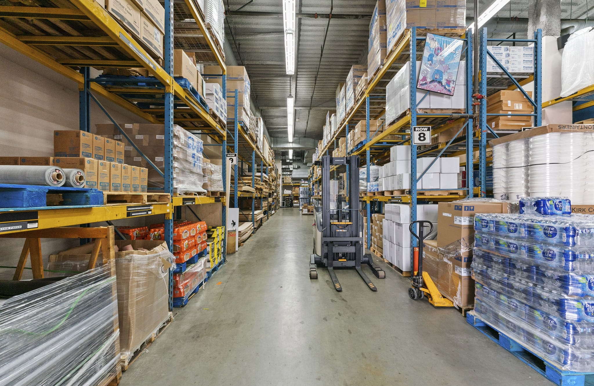 A long aisle in a warehouse with shelves stacked high with products, a forklift, and a pallet jack.