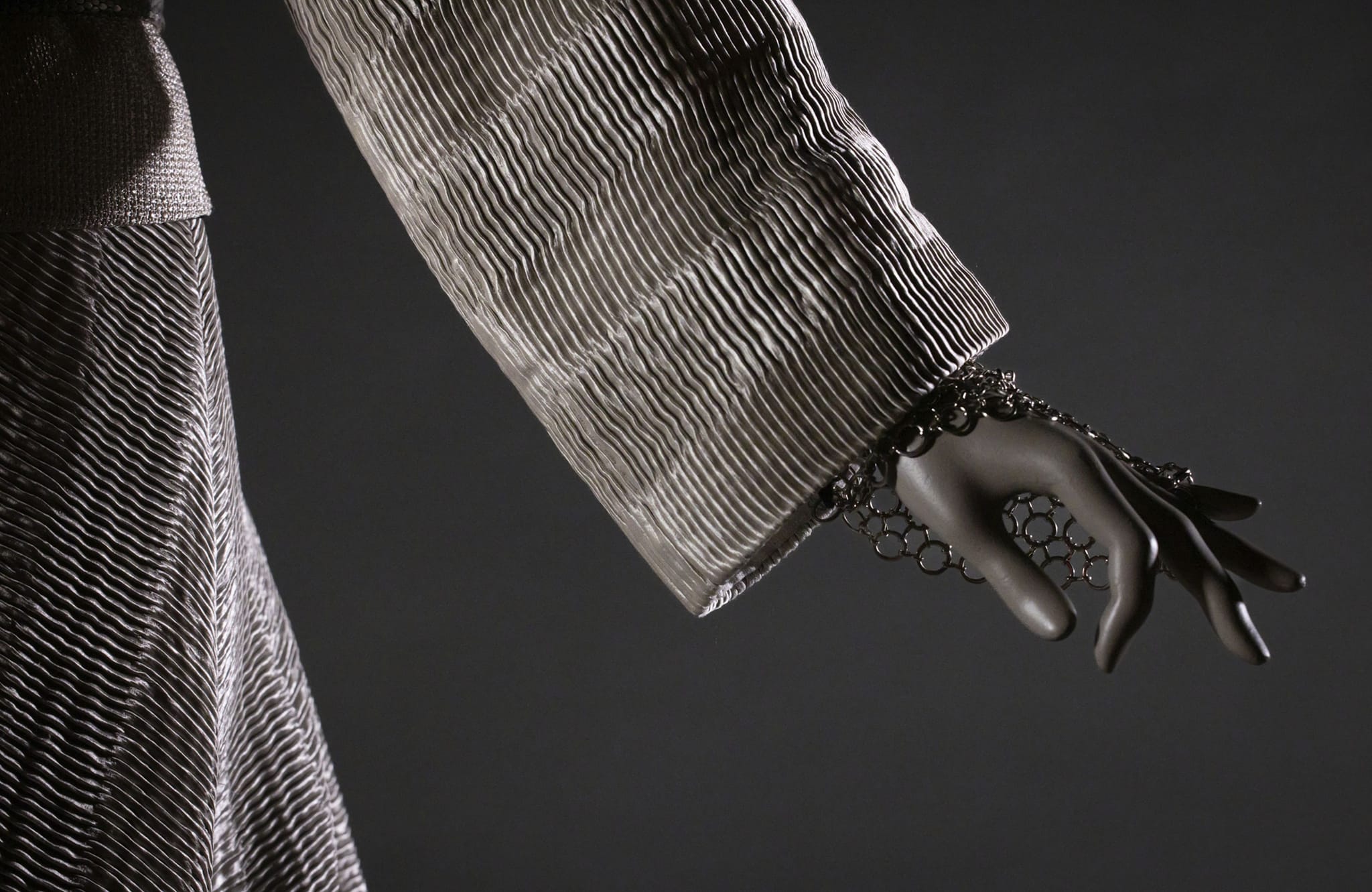 Mannequin arm in a rippled silver-gray garment with a dark metal hand chain on its fingers.