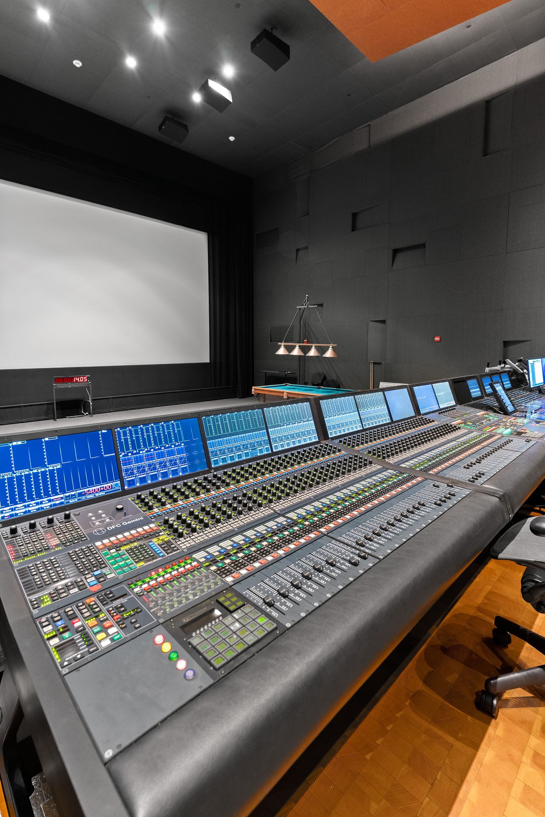 A large audio mixing console in a studio facing a blank screen, with a pool table in the background.