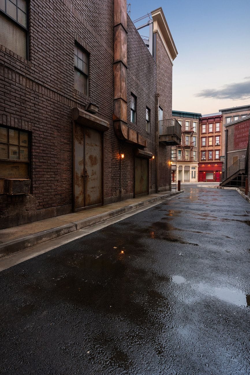 A wet brick alley with rusty doors and pipes, leading to city buildings under a pale sky.