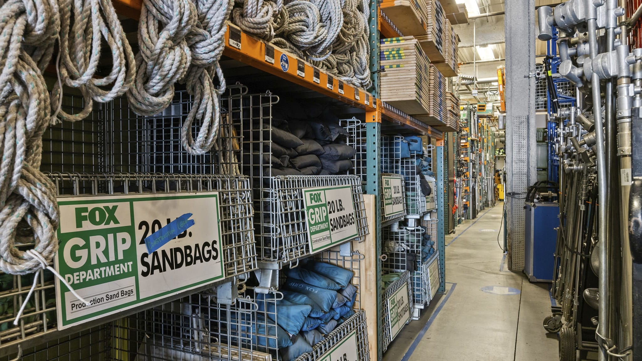 A warehouse aisle with shelves of coiled ropes and Fox Grip sandbags on the left, and film production equipment on the right.
