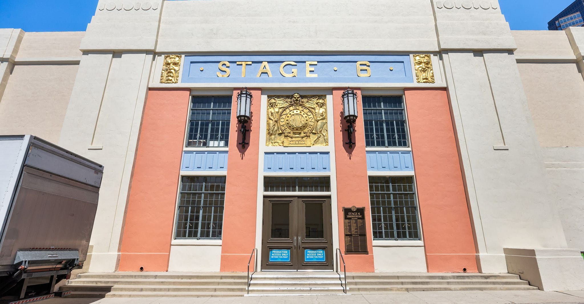 Art Deco building facade with "STAGE - 6" sign, featuring salmon, blue, and gold details, and a white truck trailer on the left.