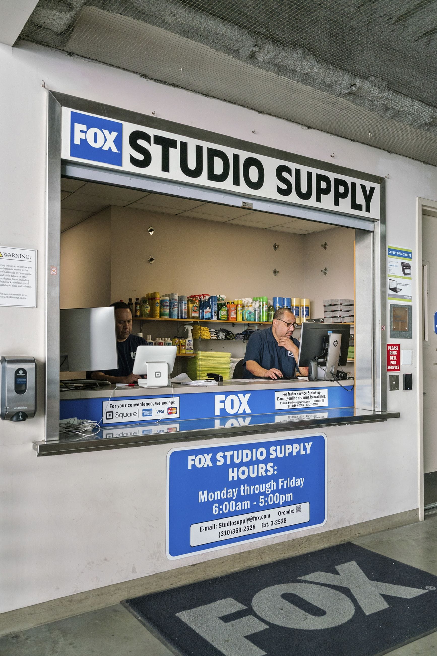 A service window for "FOX STUDIO SUPPLY" with two employees working inside, and signs displaying hours and contact information.