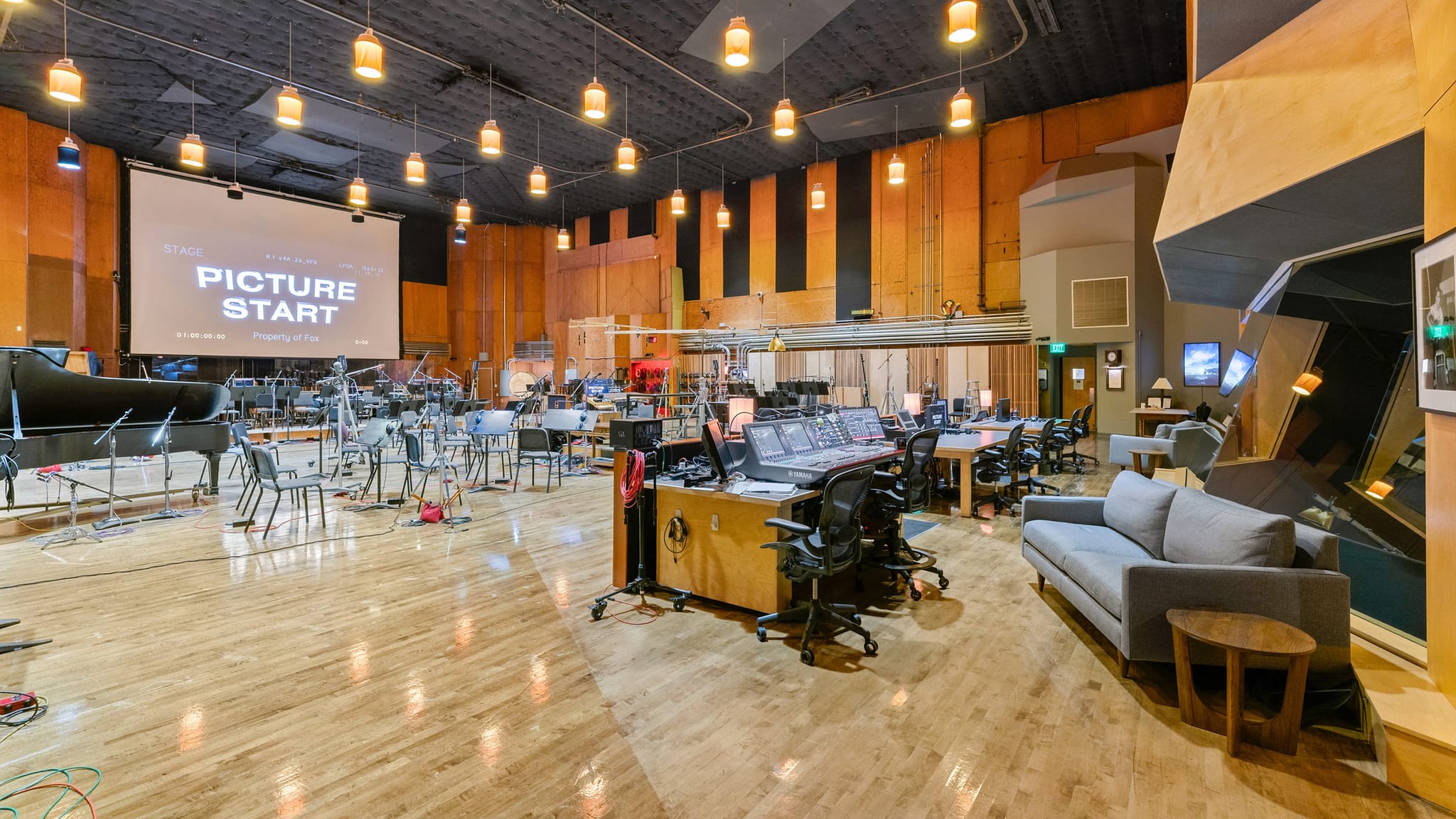A spacious recording studio with a "PICTURE START" screen, a grand piano, numerous empty music stands, and a control room with multiple workstations and a grey sofa.