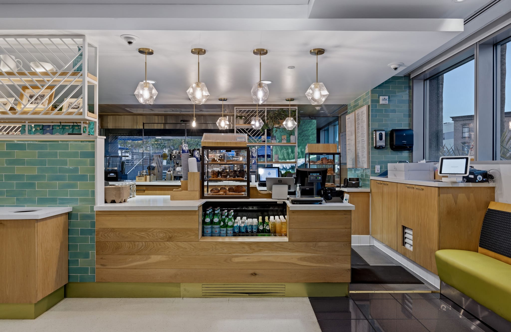 A bright, modern cafe interior with a light wood service counter, display cases of pastries, teal tiled walls, and large windows.