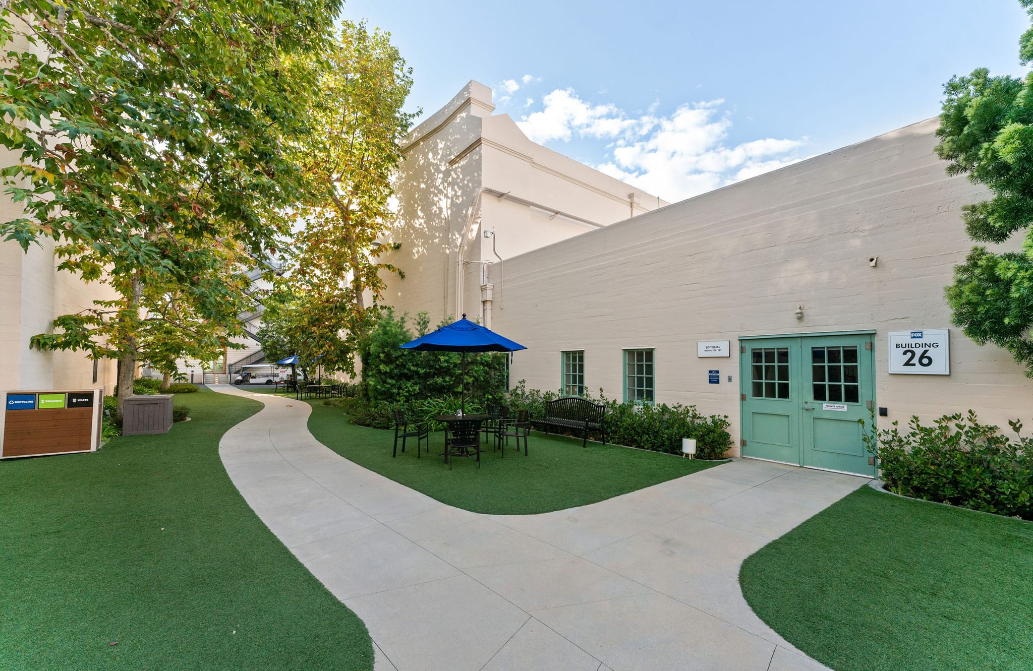 A green courtyard with a winding path leads to Building 26, featuring a large green door and an outdoor seating area with a blue umbrella.