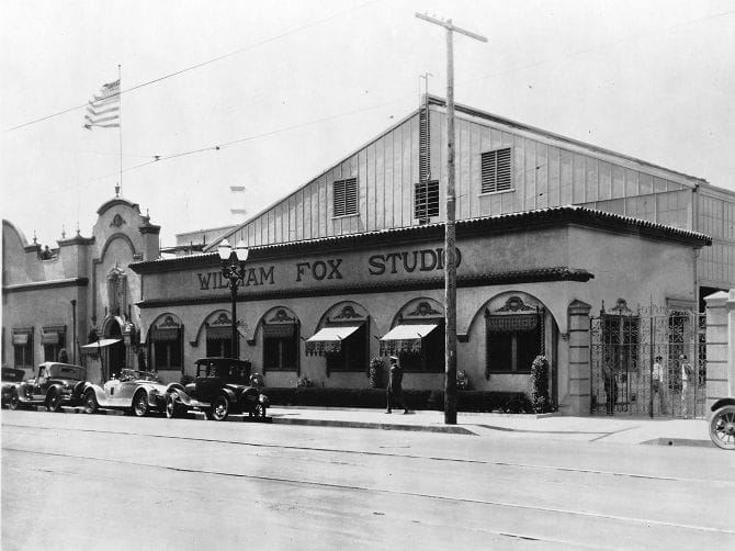 A vintage black and white photo of the William Fox Studio building, an American flag flying, and several classic cars parked outside.