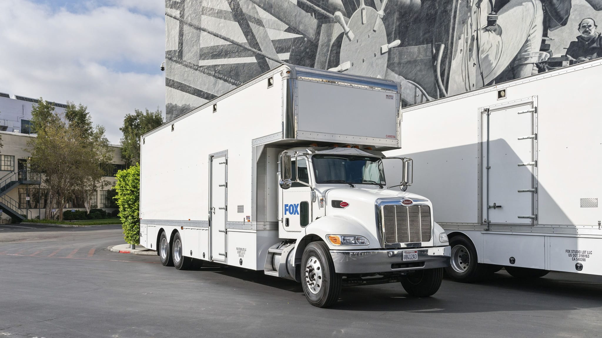 A white FOX semi-truck with a large production trailer is parked in front of a building with a grayscale mural.