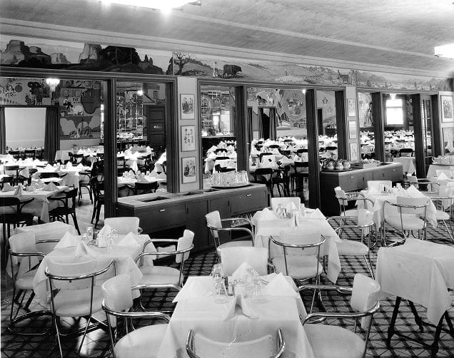 Black and white photo of an empty dining room with tables set for service and murals on the upper walls.