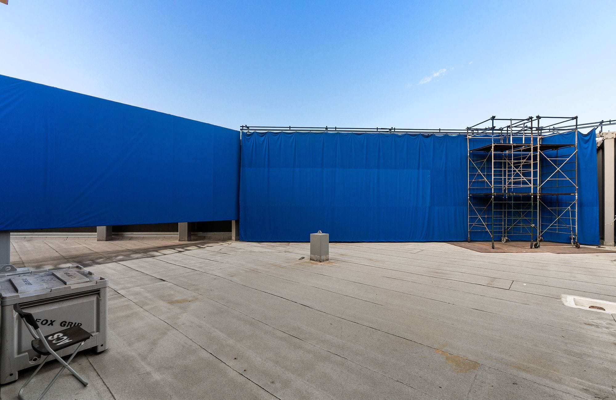 An outdoor film set with a long blue screen wall, scaffolding, and a grip box on concrete ground.