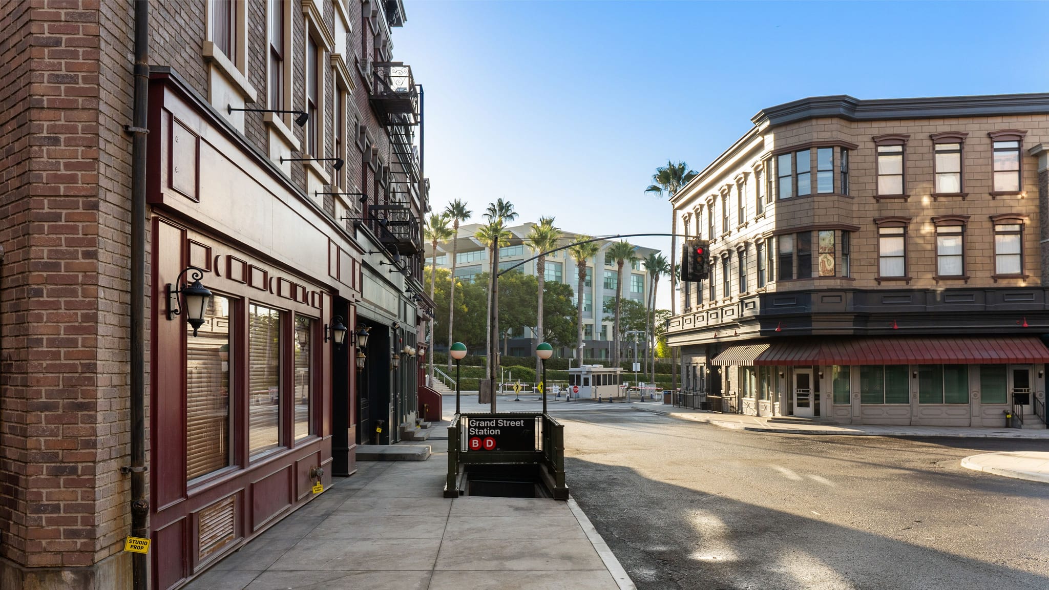 A sunny street scene featuring old-style brick buildings, a "Grand Street Station" subway entrance, and palm trees with modern buildings in the background.