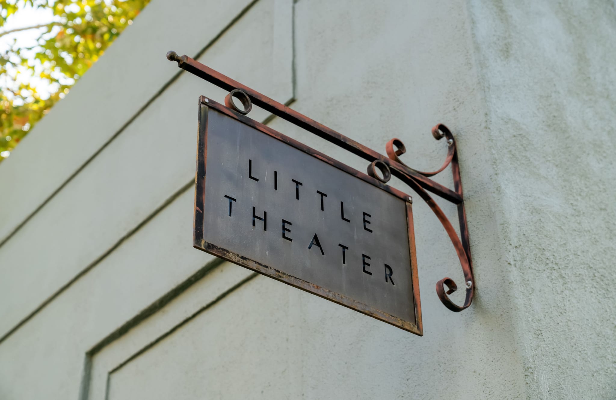 A dark metal sign for "LITTLE THEATER" hangs from a decorative wrought iron bracket on a light-colored building.