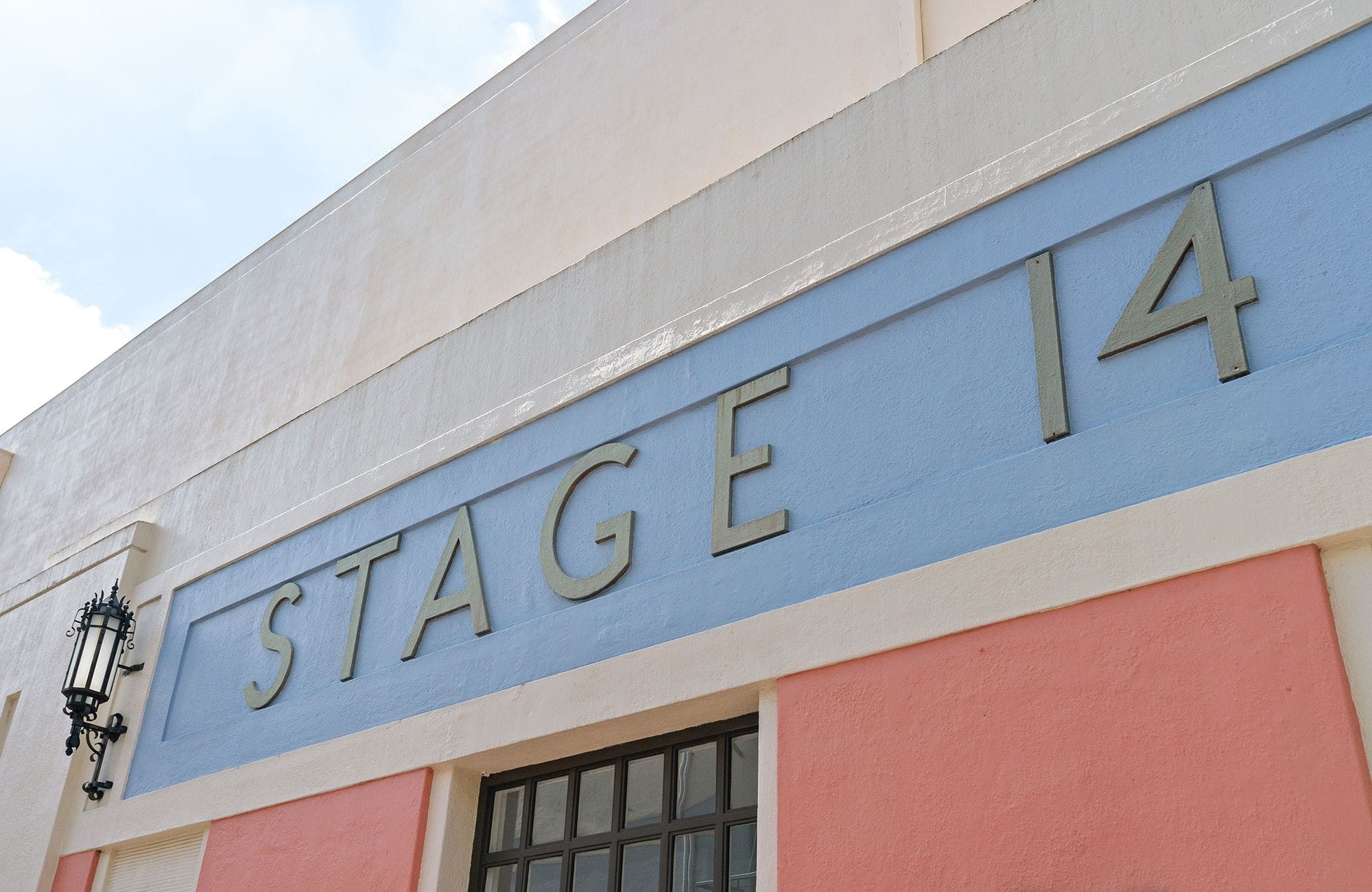 Exterior of a building with "STAGE 14" in raised letters on a blue band, set against cream and peach walls.