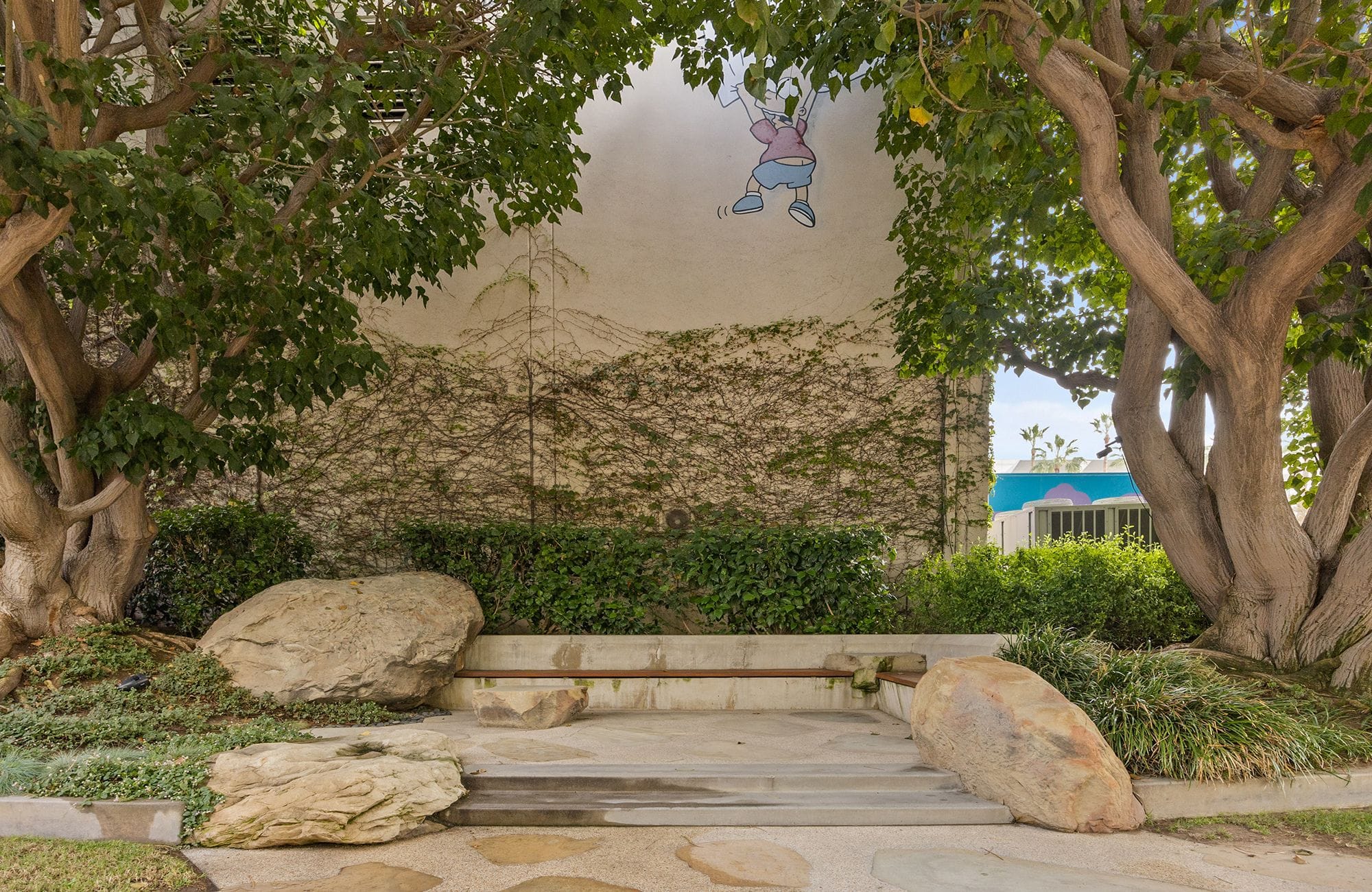 A wall with ivy and a cartoon of a child, framed by two trees and landscaping with large rocks.